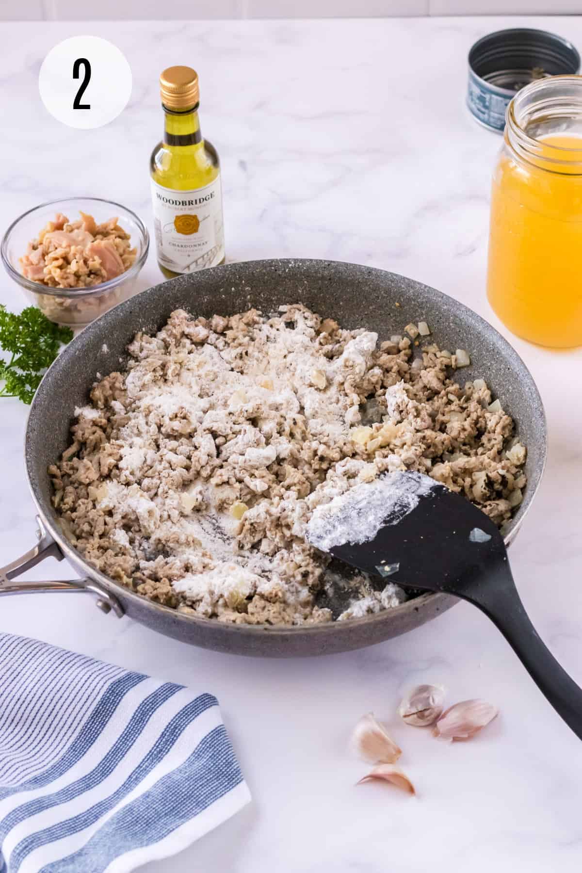 Browned ground turkey mixture with flour in skillet with chopped clams, wine and chicken broth in upper background with  garlic cloves below and blue & white towel in lower left. 