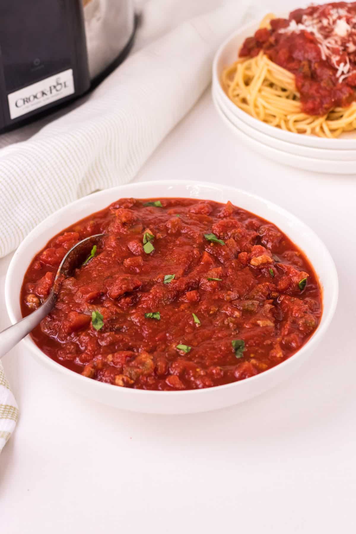 White bowl with spaghetti meat sauce and plates of spaghetti and sauce  and slow cooker in upper background.