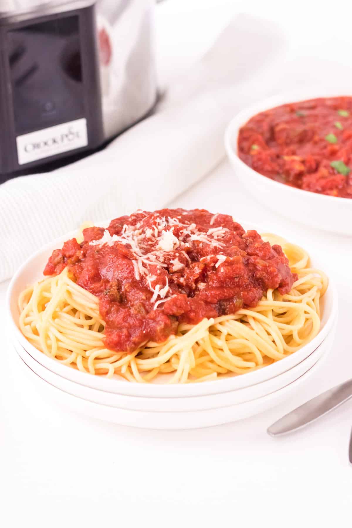 Stack of white plates with spaghetti and meat sauce topped with shredded cheese with bowl of sauce and slow cooker in background.
