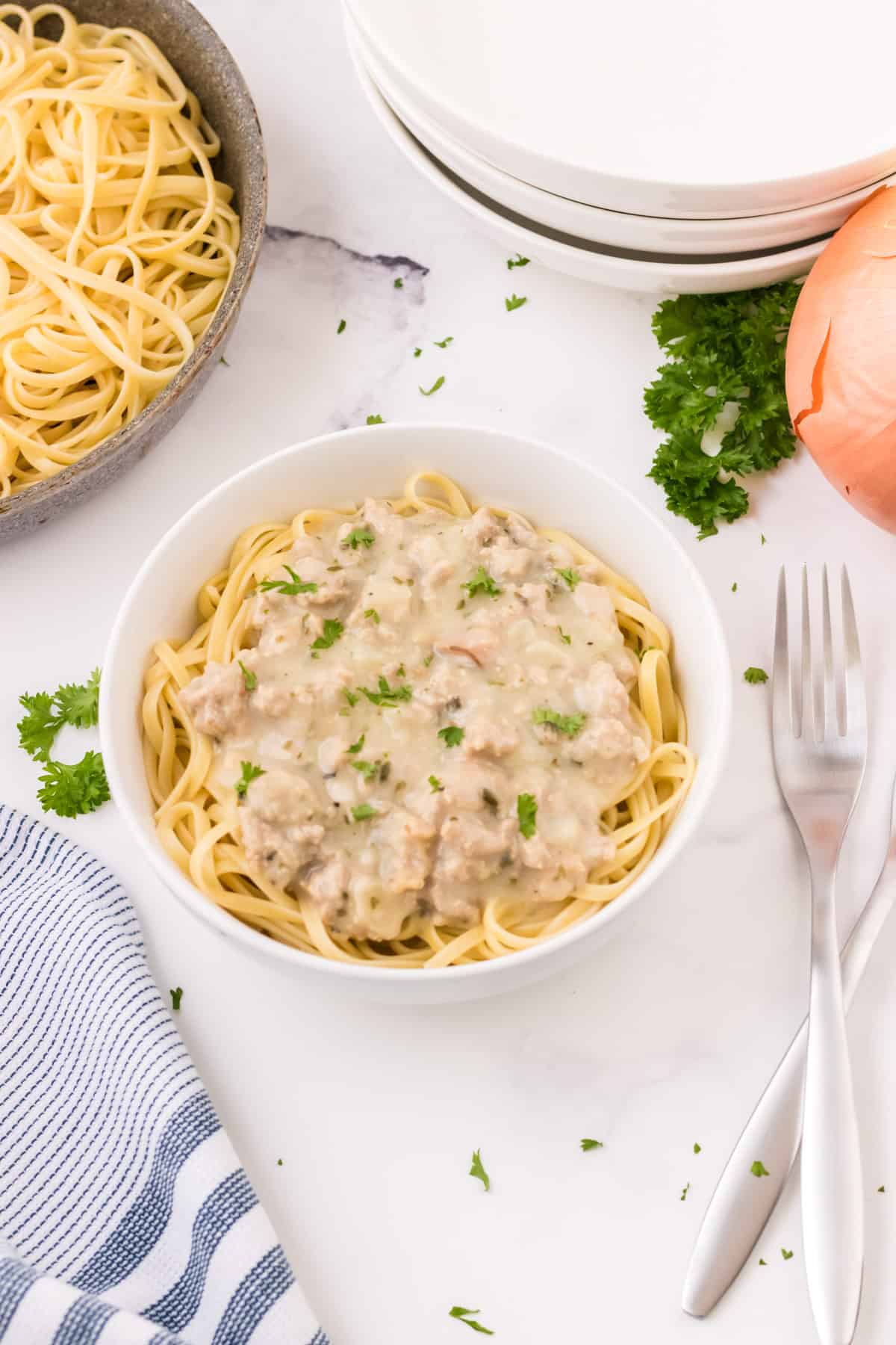 Top view image of creamy ground turkey and clam sauce with linguine in white bowl with chopped parsley on top and skillet of pasta, white bowls, sprig of parsley and onion in upper background with forks and blue and white towel in lower background.