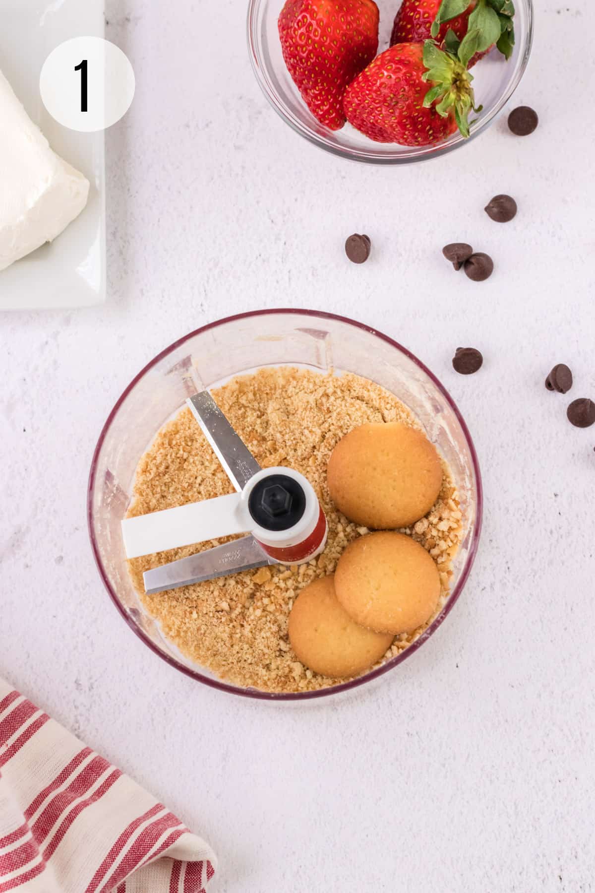 Handheld food chopper with vanilla wafer cookies finely chopped and chocolate chips, strawberries and cream cheese in upper background. 