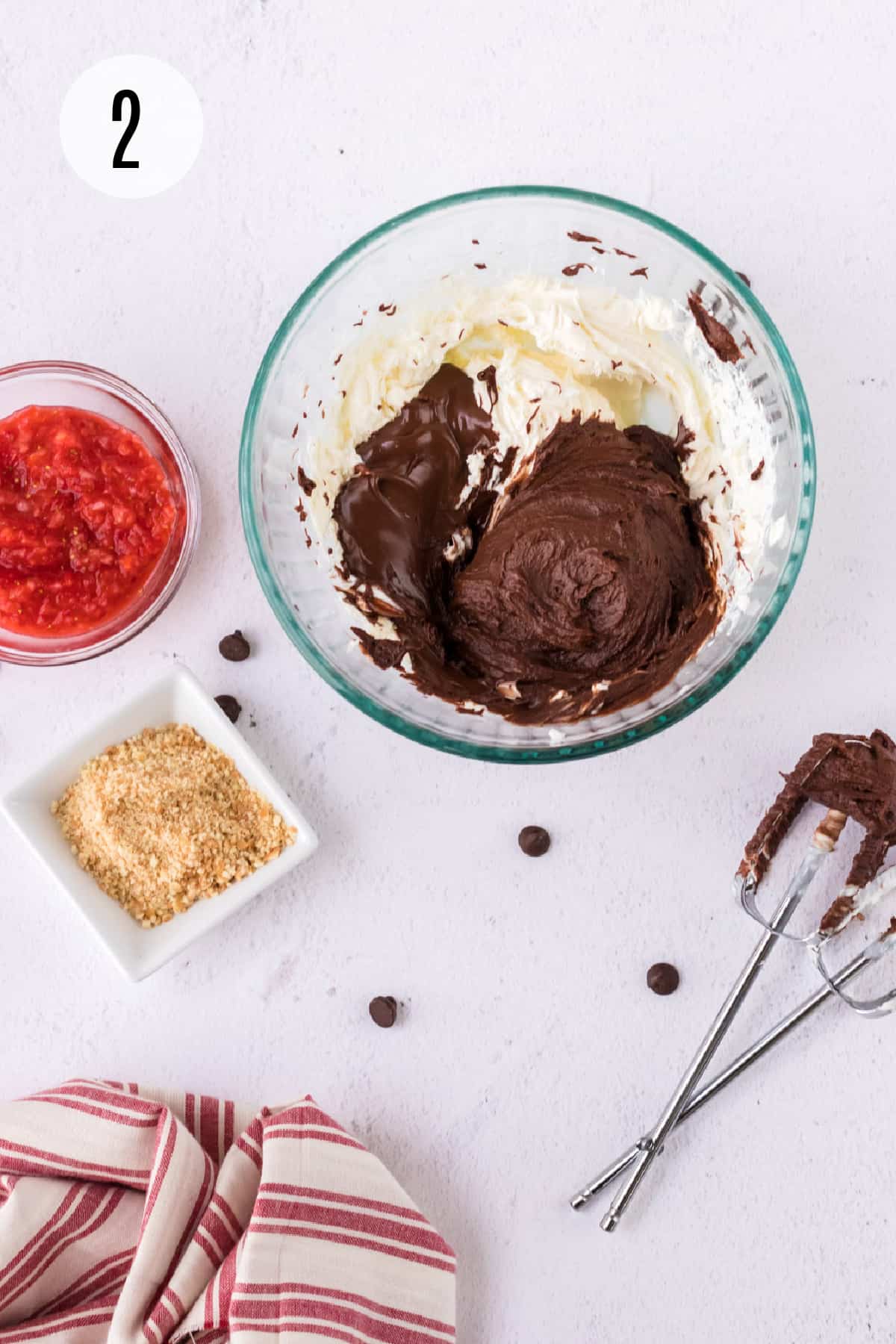 Glass bowl with melted chocolate and cream cheese blended and small bowls of pureed strawberries, crushed vanilla wafers and mixer beaters surrounding bowl. 