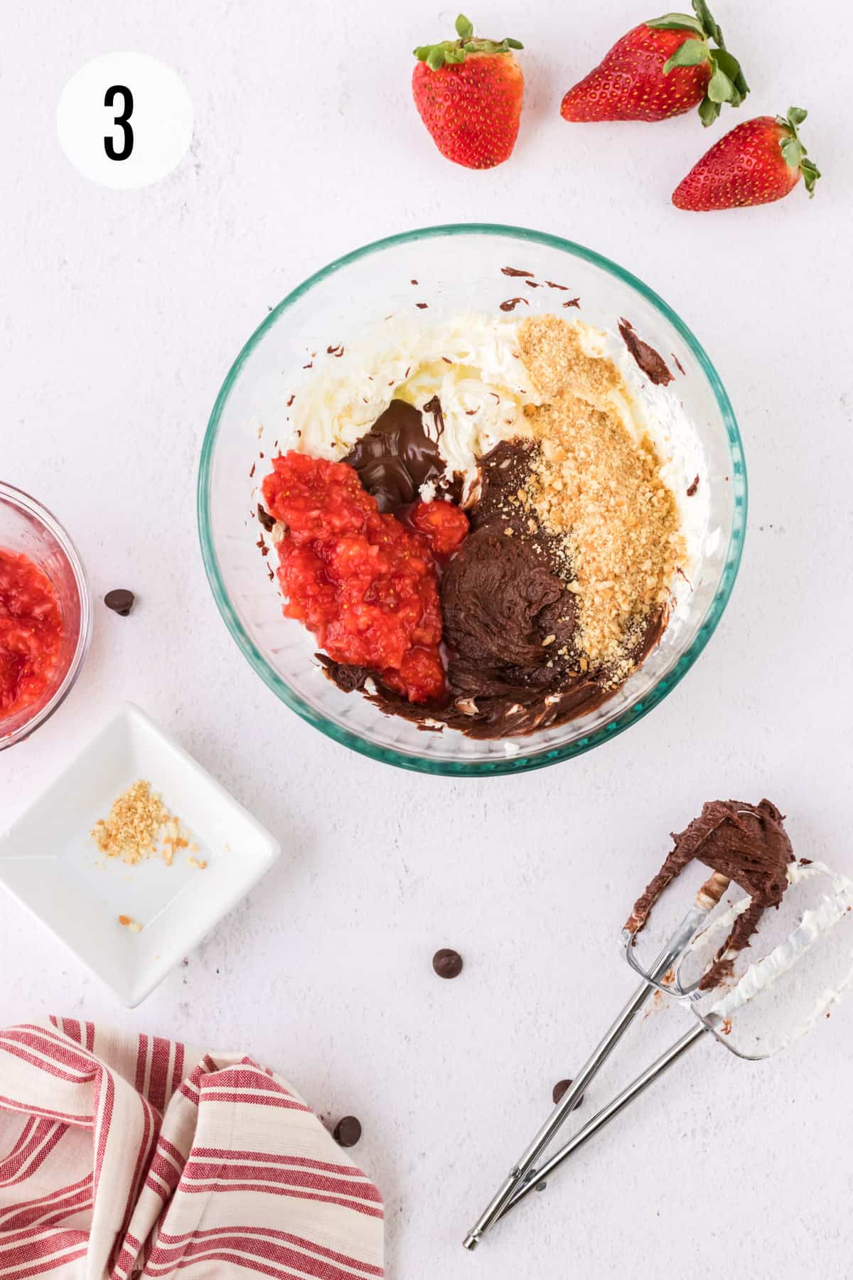 Glass bowl with pureed strawberries, melted chocolate, vanilla wafer crumbs and cream cheese with whole strawberries and mixer beaters surrounding bowl. 