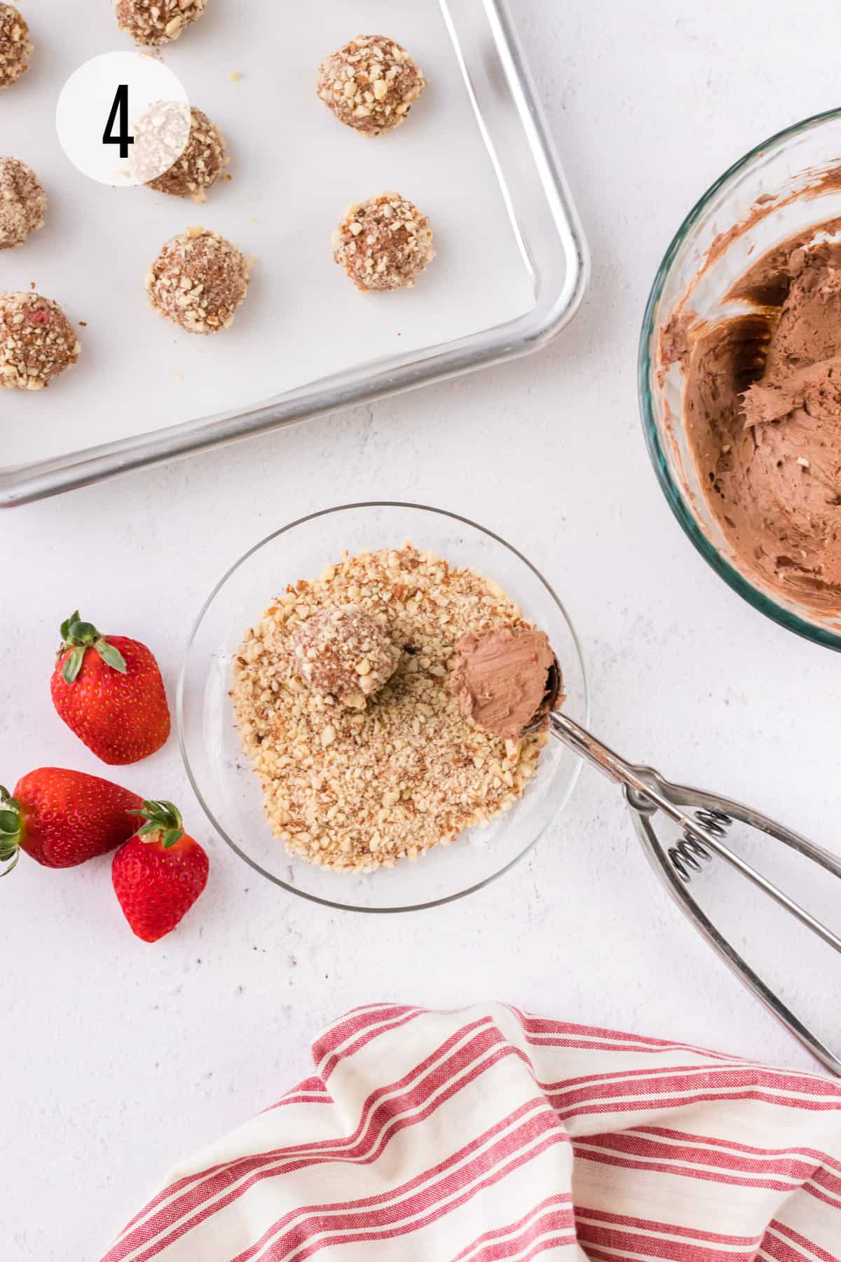 Bowl of finely chopped almonds with silver scoop rolling cheesecake bites with whole strawberries, tray of bites and bowl of cheesecake mixture and red and white linen surrounding. 