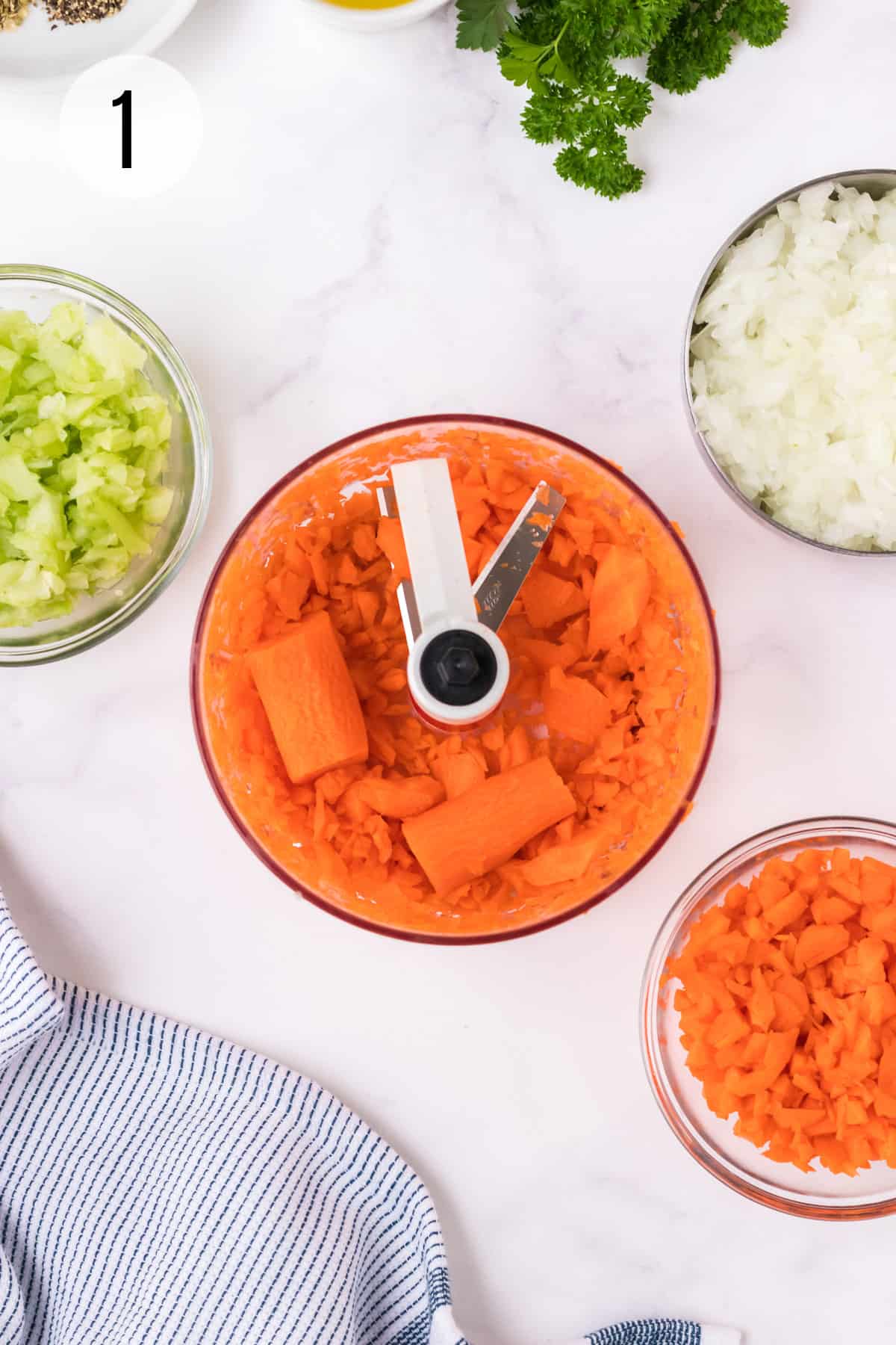 Handheld food chopper with chopped carrots and bowls of chopped celery, onions and more carrots around with blue & white towel in lower left. 