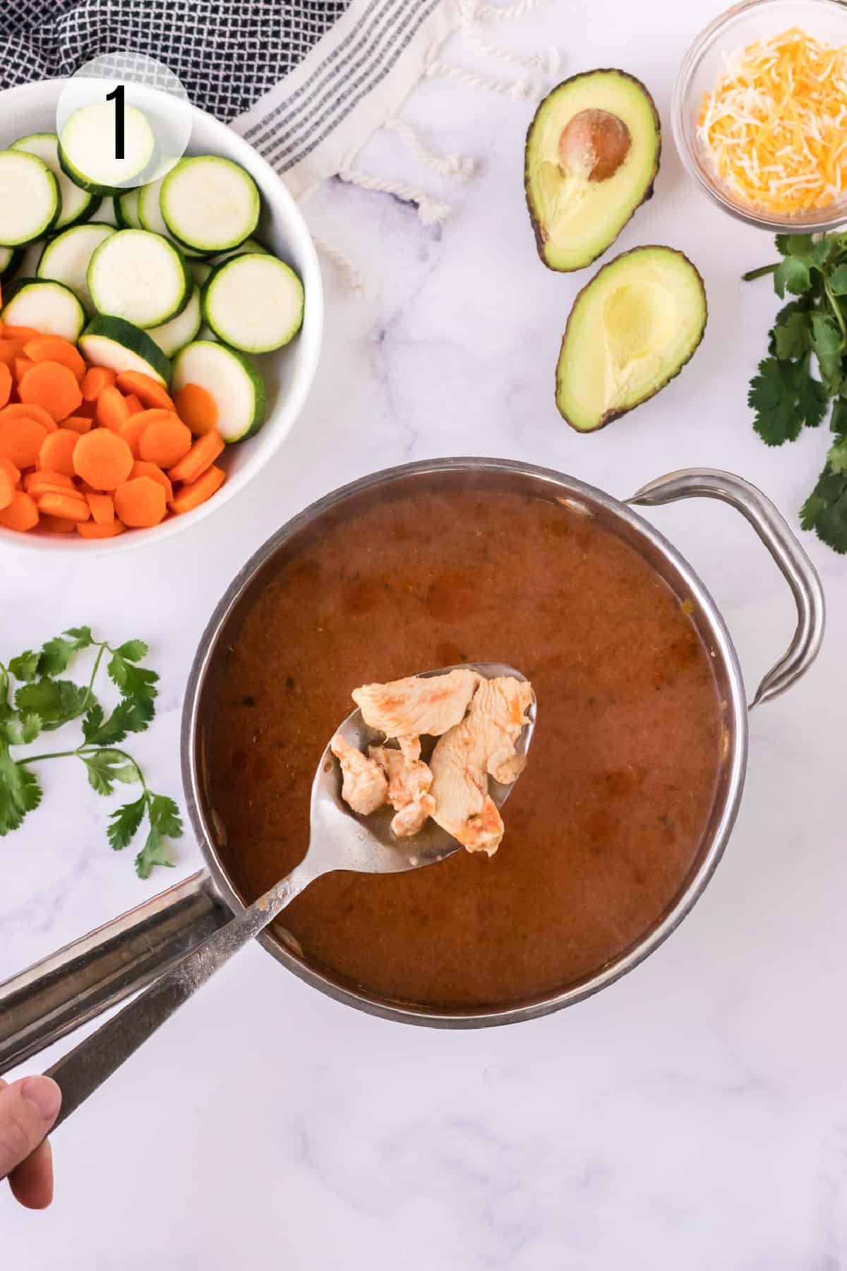 Pan of cooked tortilla soup broth with cooked chicken and bowl of sliced carrots and zucchini and avocados and shredded cheese.