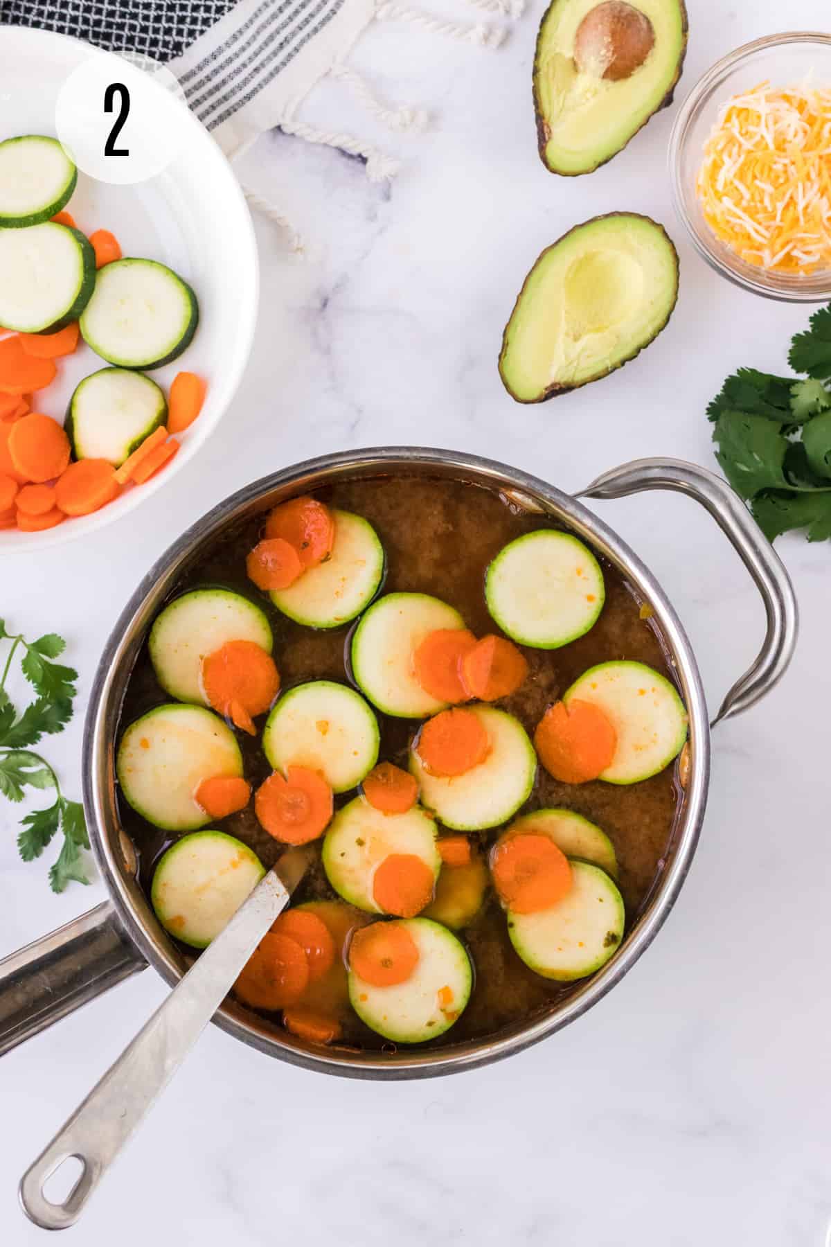 Pan of chicken tortilla soup with sliced zucchini and carrots with bowl of veggies and avocado, cheese and cilantro in upper background.