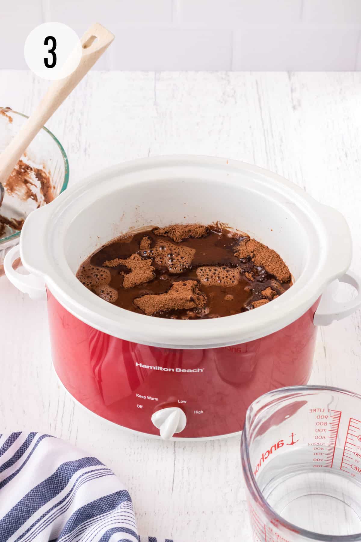 Red and white slow cooker with chocolate lava cake mixture inside and glass measuring cup in lower right, glass batter bowl with spoon in upper left and blue and white linen in lower left corner. 