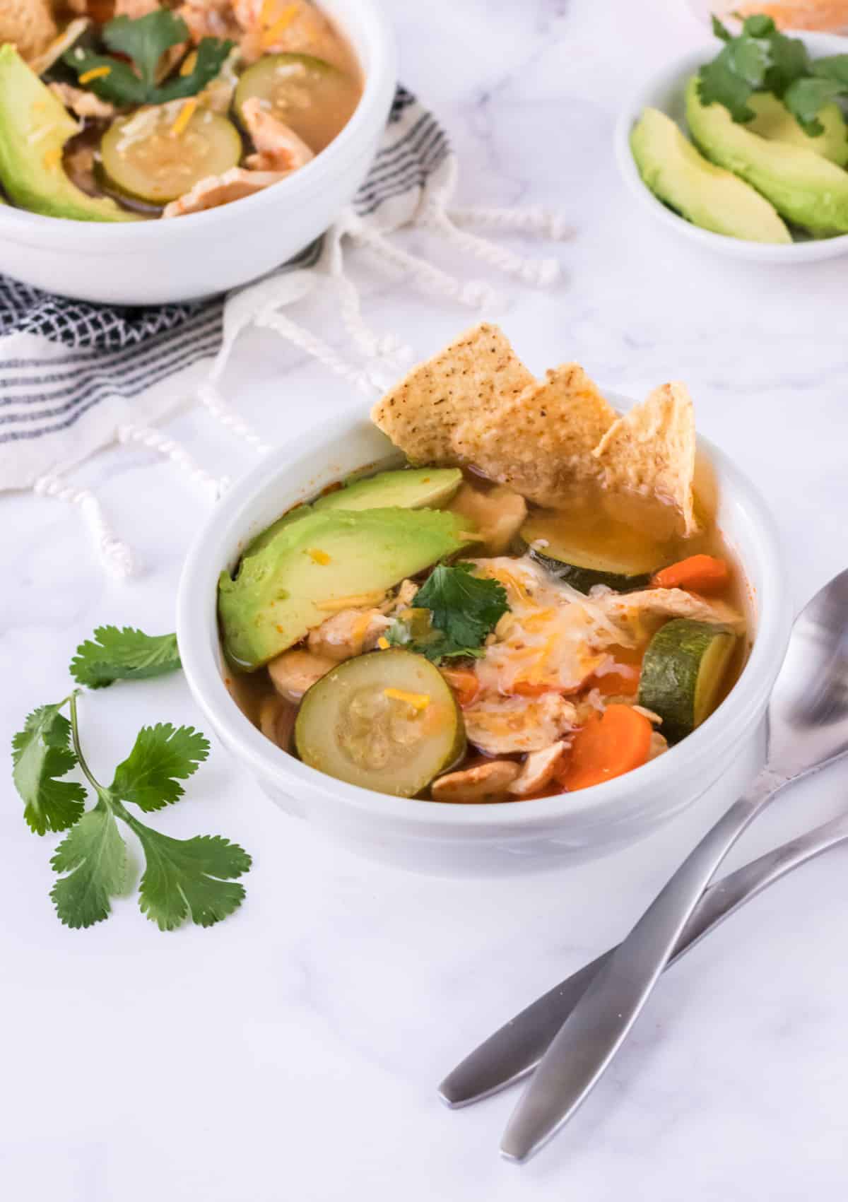 Bowl of chicken and vegetable tortilla soup with extra bowl in upper left and bowl of sliced avocados in upper right.