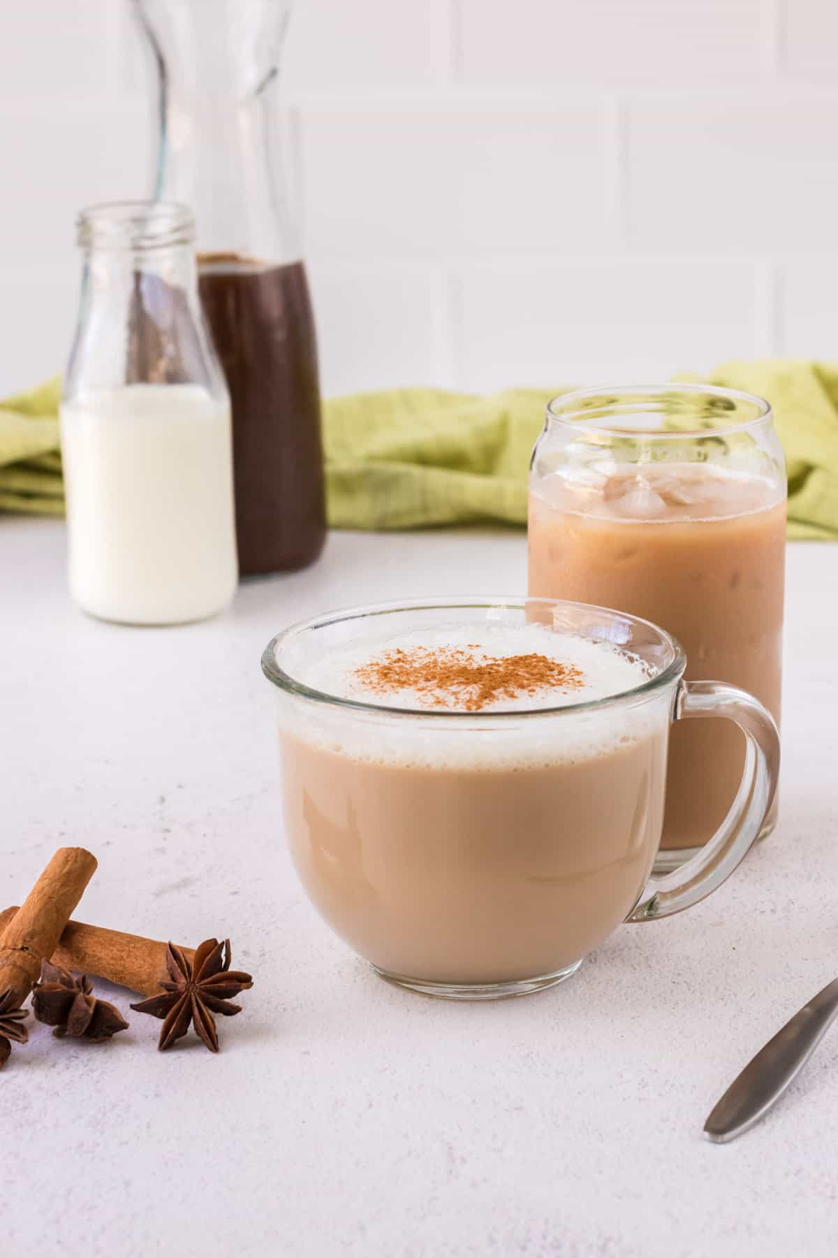 Glass mug with warm chai latte, taller glass with iced chai latte and milk and chai concentrate in background with cinnamon sticks and star anise in foreground.