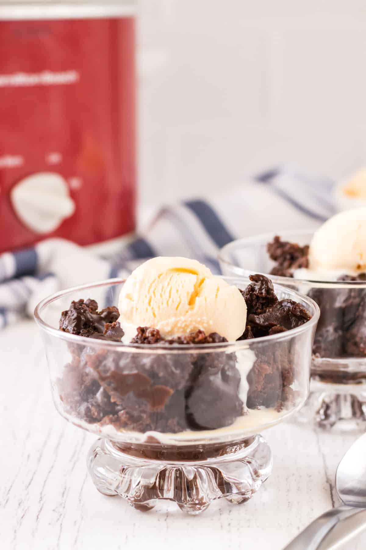 Small glass dish with chocolate pudding cake topped with vanilla ice creamand second bowl, red slow cooker and blue and white linen in background.