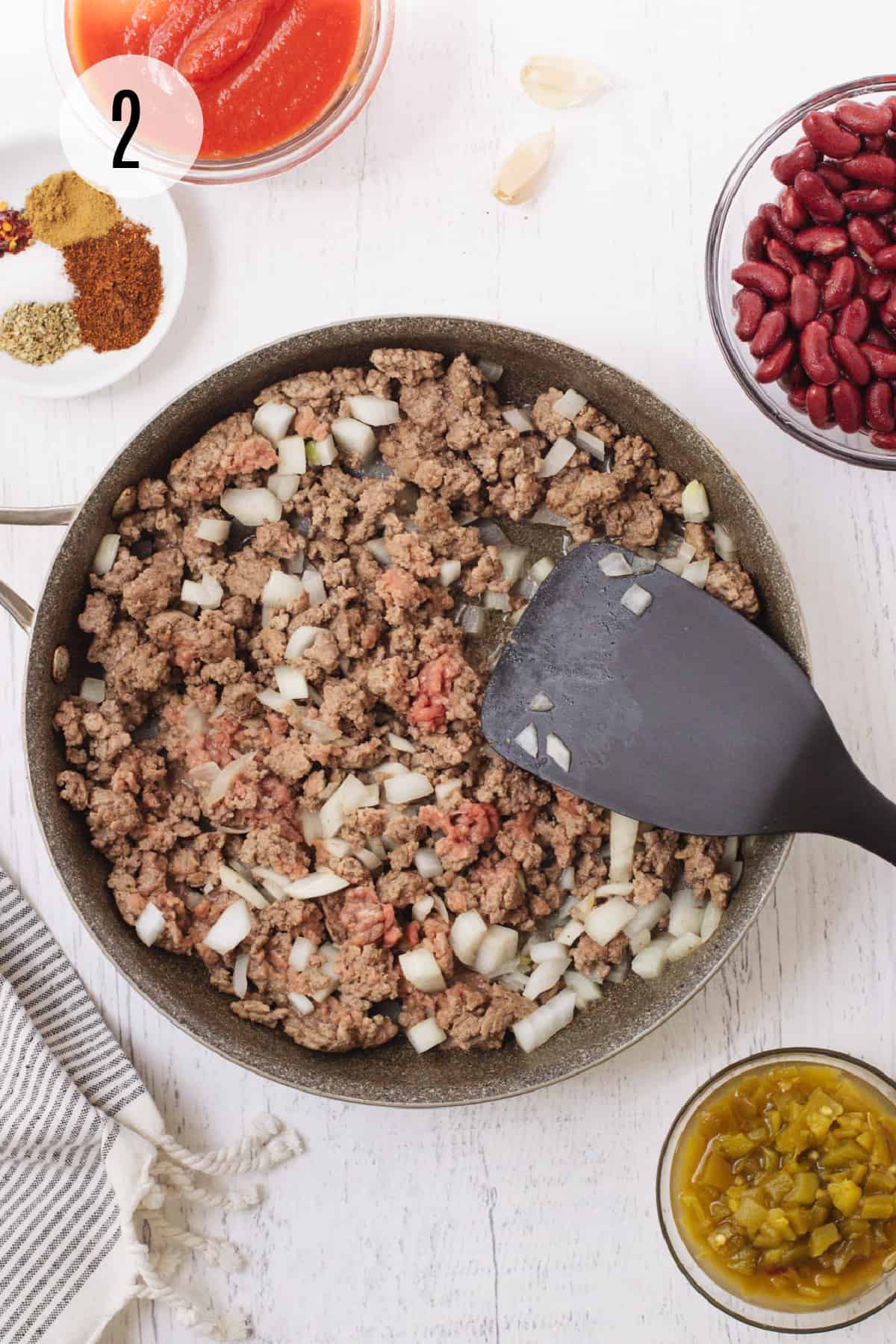 Sauté pan with ground beef and onions browning with black spatula and remaining ingredients for taco quinoa surrounding the pan. 