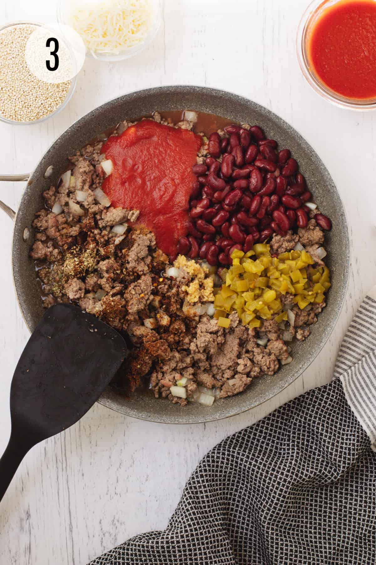 Saute pan with ingredients for ground beef and quinoa taco skillet including kidney beans, green chilies, tomato sauce and spices with black spatula and black and white linen in foreground and quinoa and shredded cheese in upper background. 