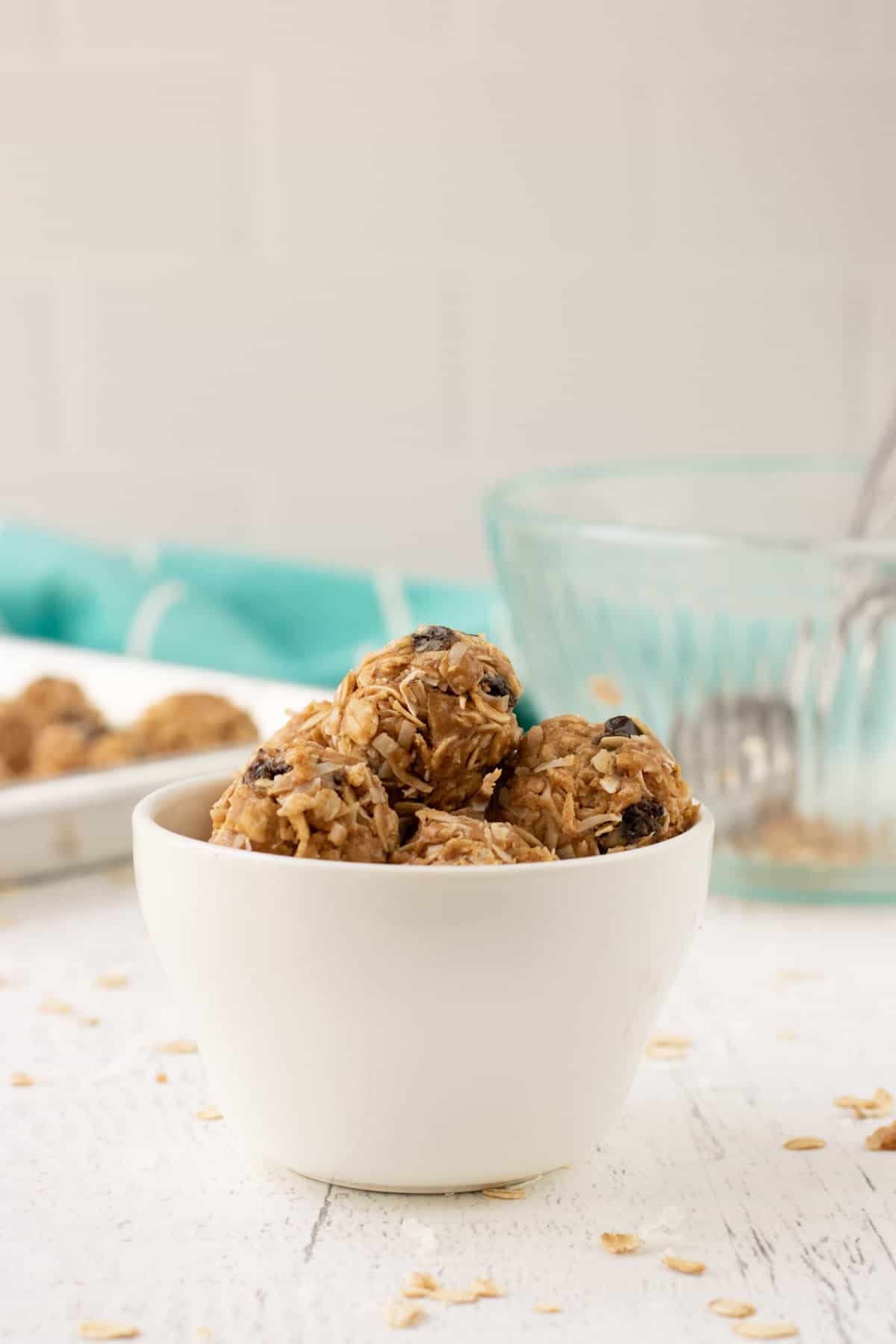 No Bake Peanut Butter Oatmeal Balls in a white bowl with tray of balls and empty ingredient bowl and aqua linen in background.