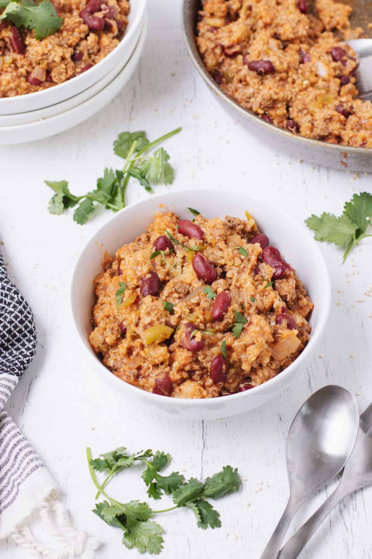 White bowl of taco spiced ground beef and quinoa with kidney beans with cilantro as garnish and extra in upper background and black and white linen and spoons in foreground.