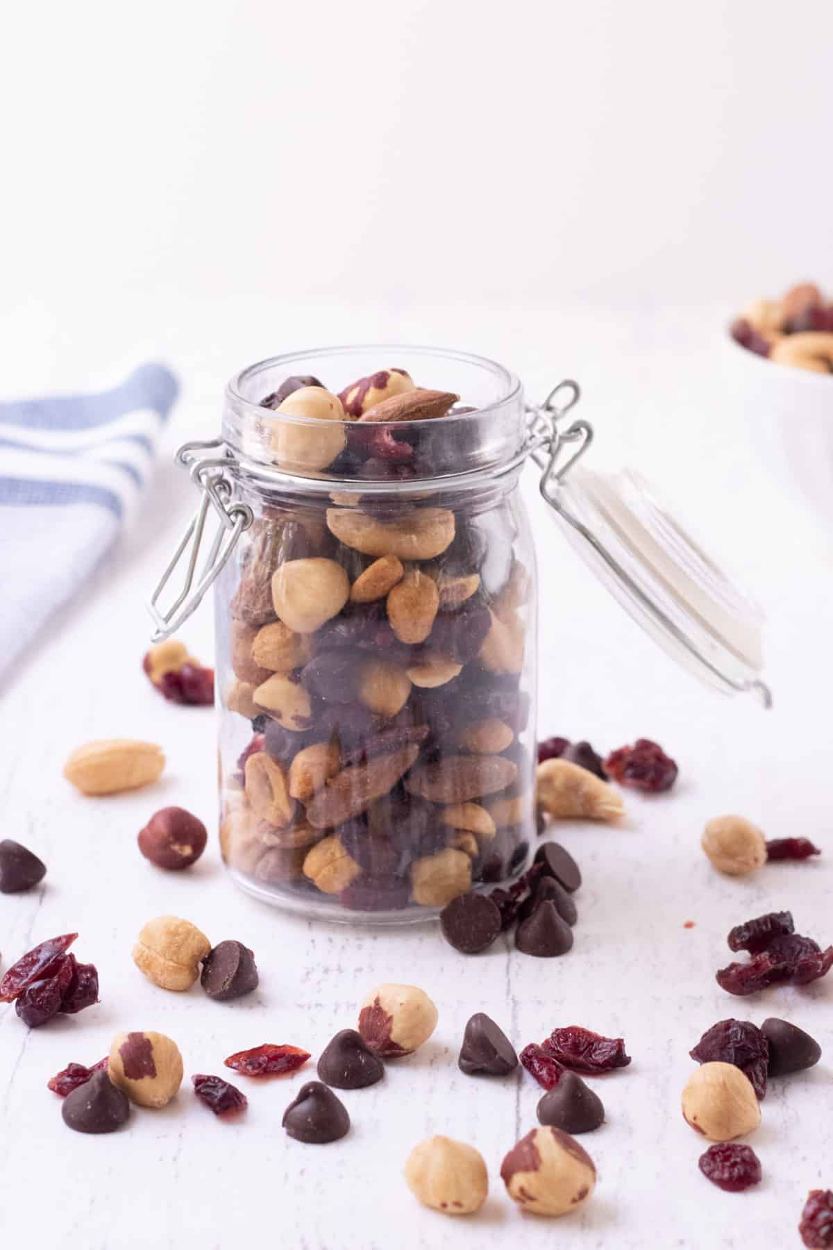 Glass jar with open lid and cherry and chocolate trail mix inside and sprinkled around with blue and white towel in upper left background.