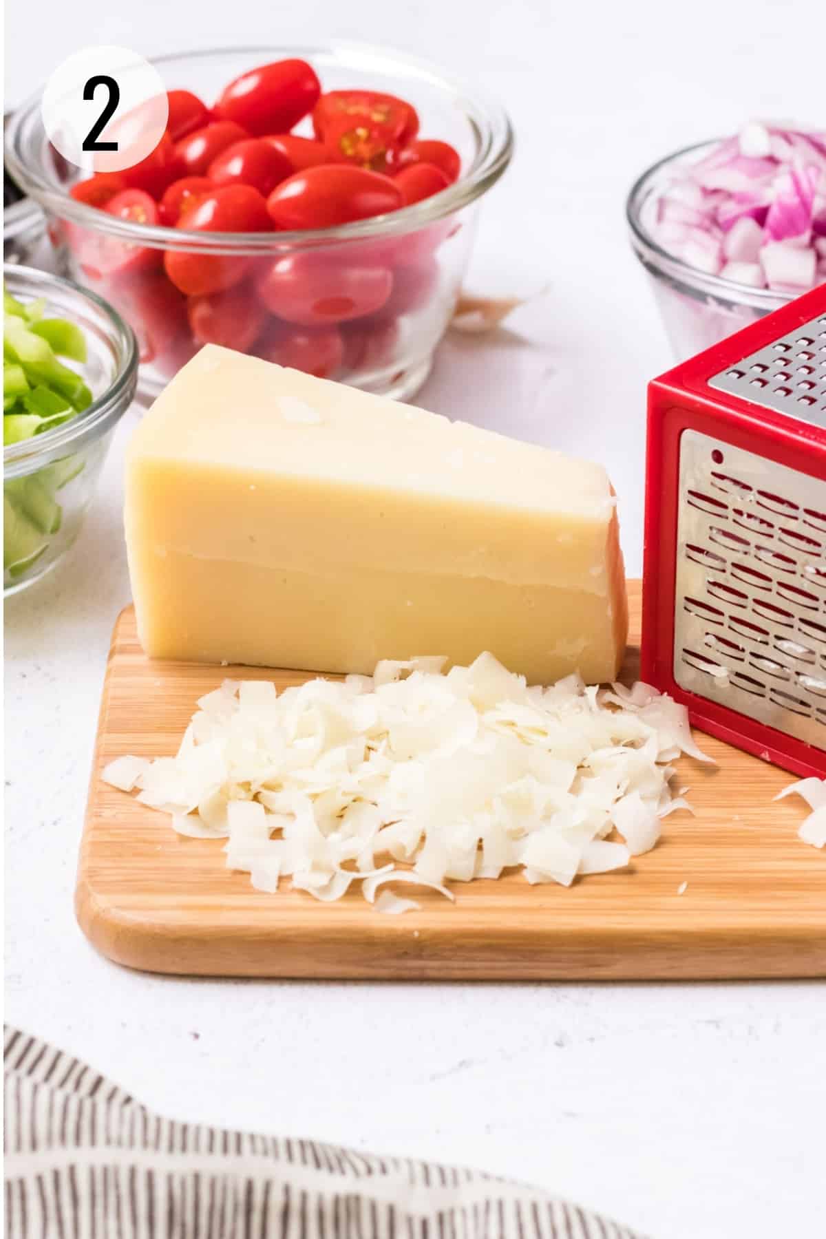 Piece of Parmesan cheese with shredded cheese using a red and silver box grater and bowls of tomatoes, green bell pepper and red onions surrounding. 