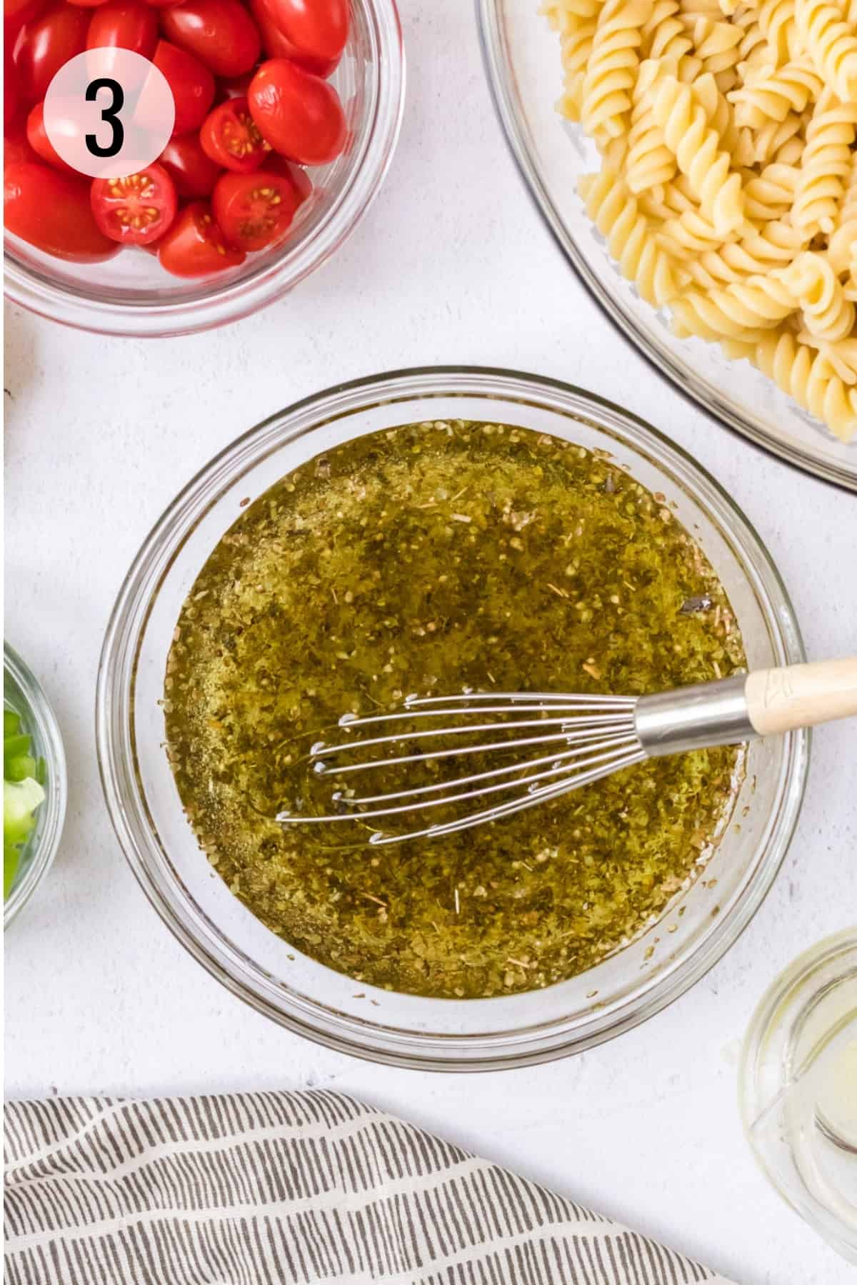 Glass bowl of olive oil, vinegar and dried herbs stirring with metal whisk and bowls of grape tomatoes and cooked pasta in upper background.