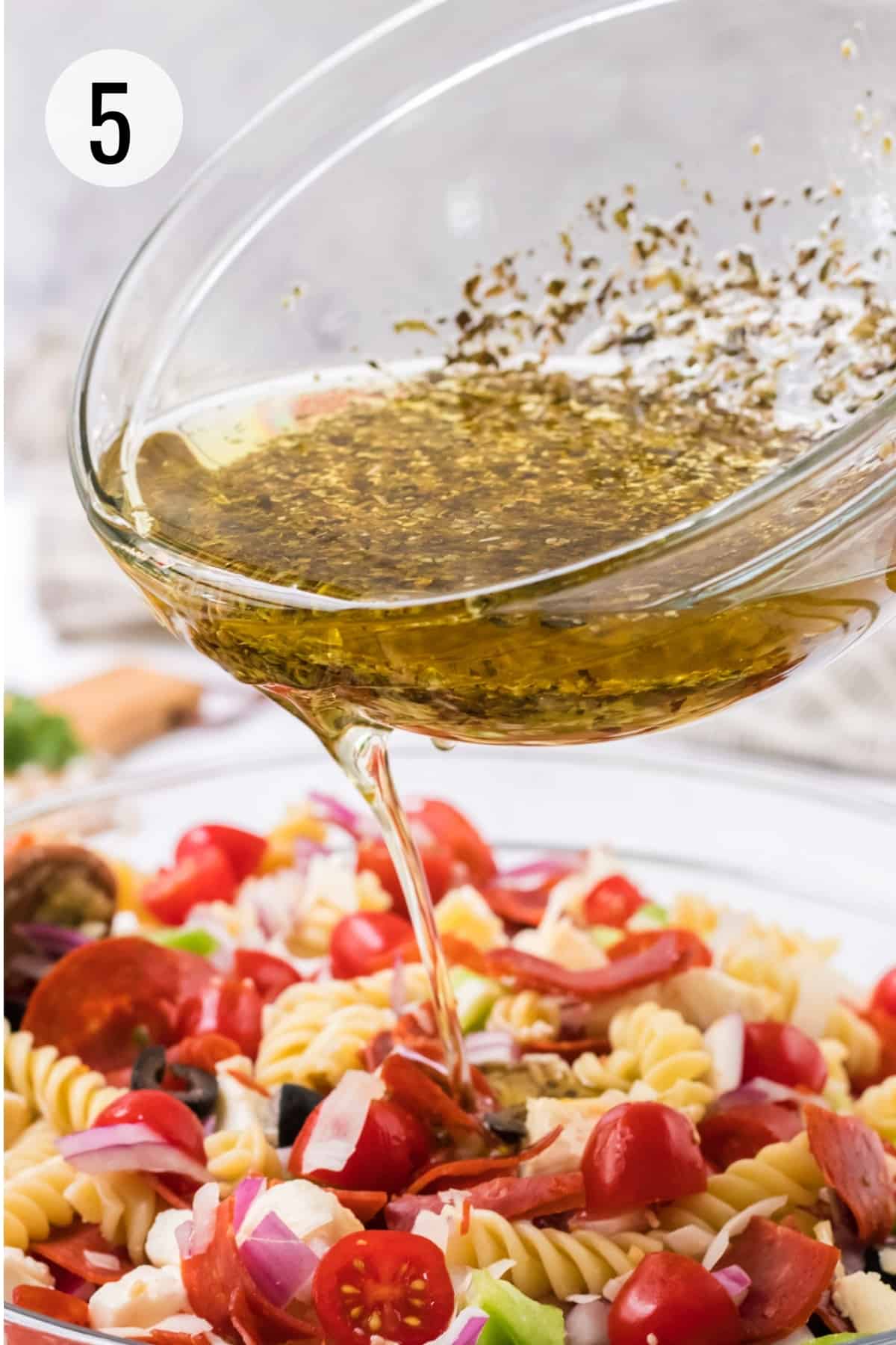 Glass bowl pouring Italian herb oil and vinegar dressing on pizza pasta salad with pepperoni.