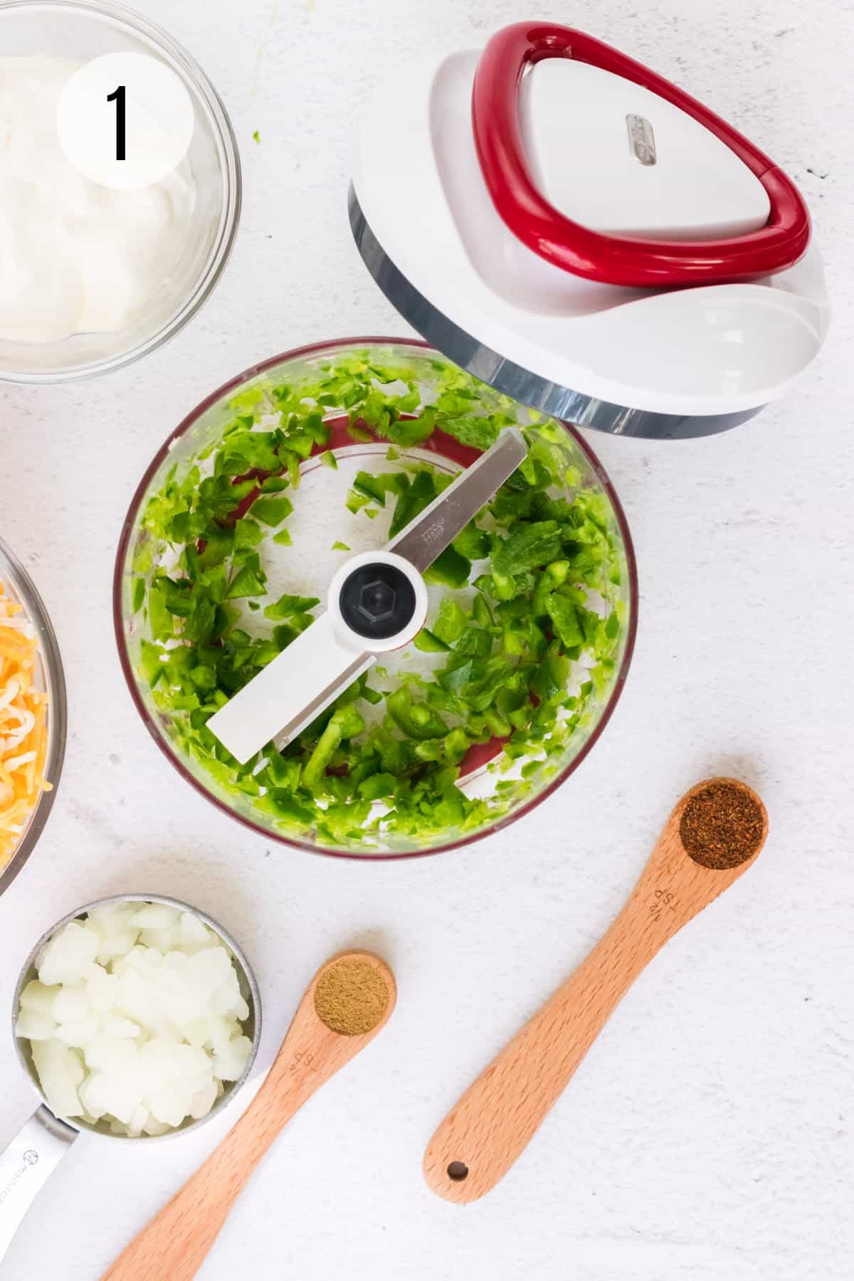 Red and white handheld food chopper with diced jalapenos inside and chopped onions, cumin and chili powder measuring cup and spoons in lower foreground.