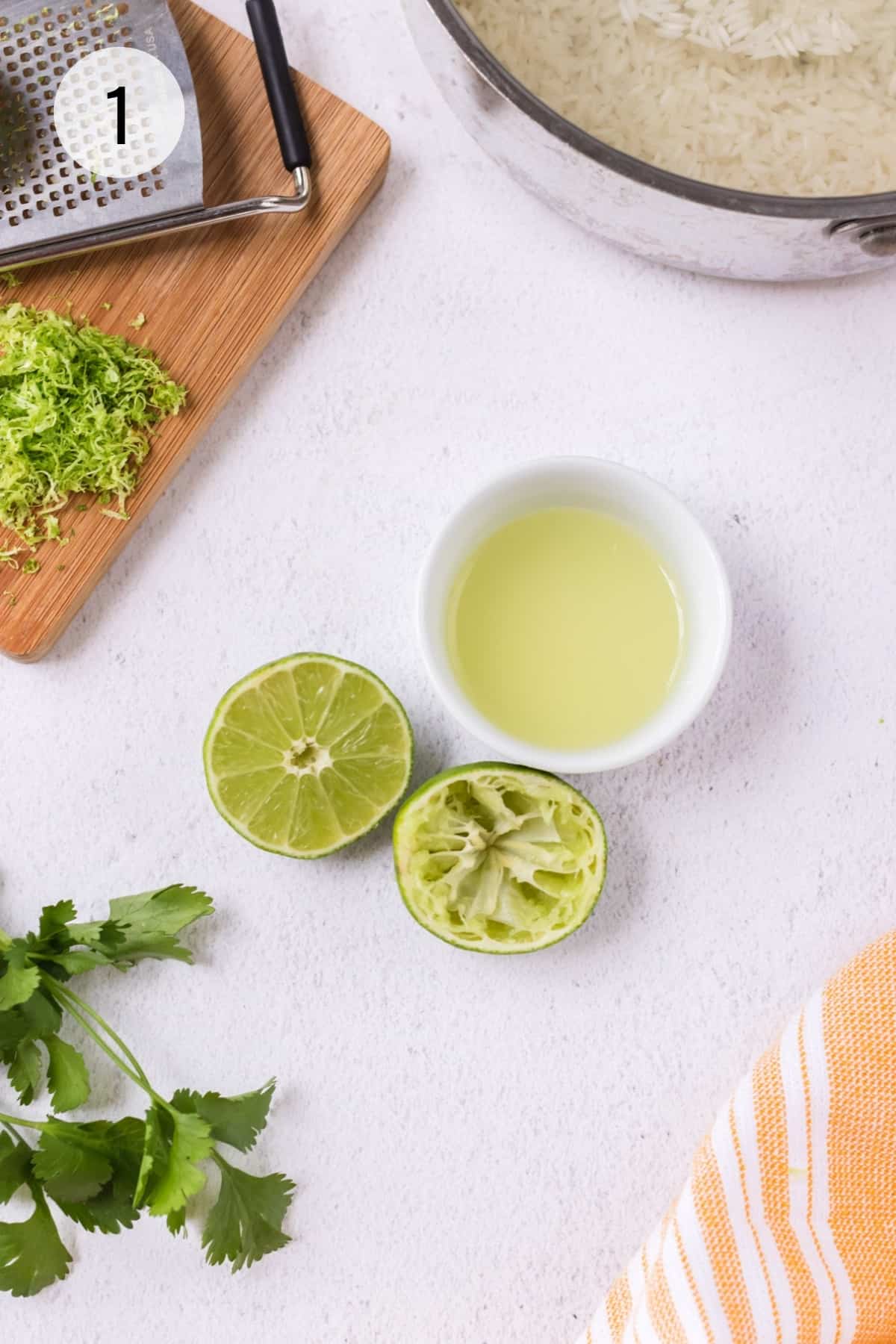 Bowl of fresh lime juice with cut limes adjacent, one squeezed and cutting board with lime zest and zester and silver pan of rice in upper background.