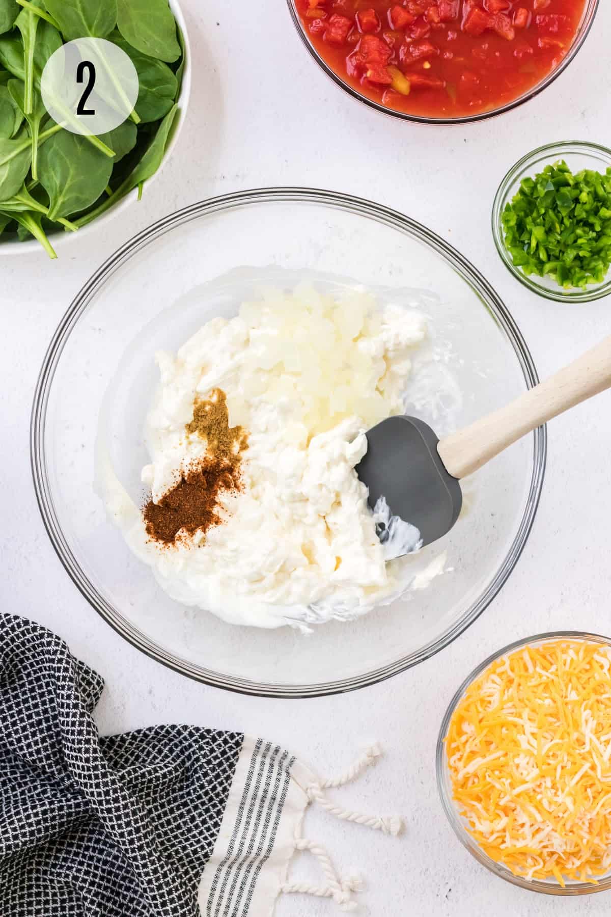 Glass bowl with cream cheese, sour cream, chopped onions and chili powder and cumin stirred by grey spatula with queso ingredients in bowls surrounding.