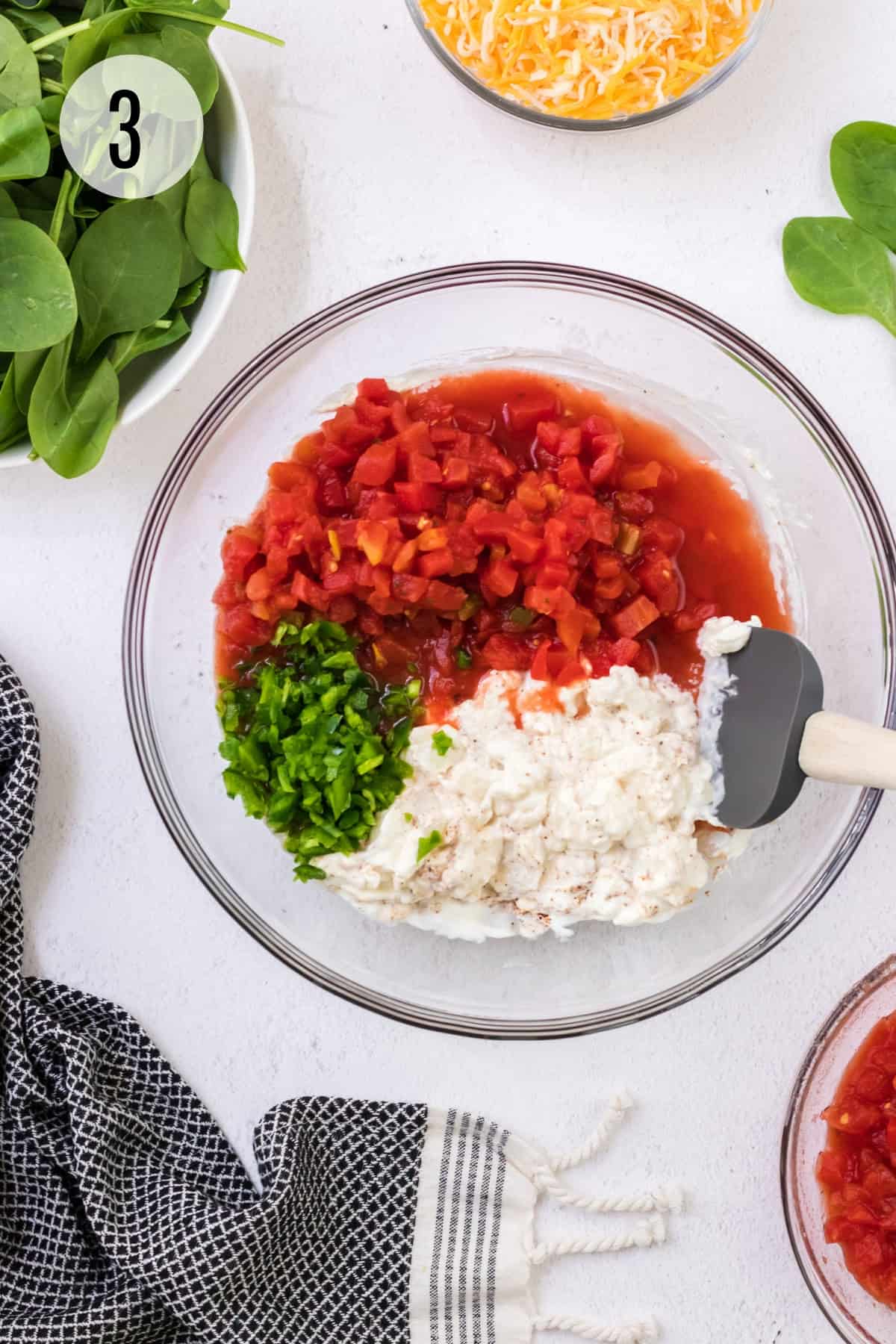 Cream cheese mixture with chopped jalapenos and diced tomatoes in large glass bowl with grey rubber spatula stirring and bowls of spinach and cheese in upper background.