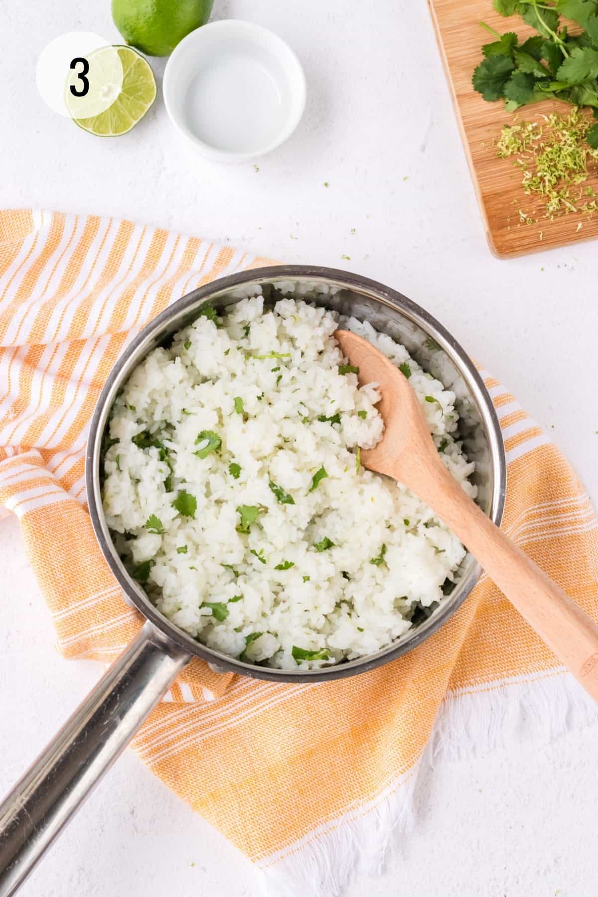Metal saucepan with white rice and chopped cilantro with wooden spoon and orange linen and limes, zest and cilantro sprigs in upper background.