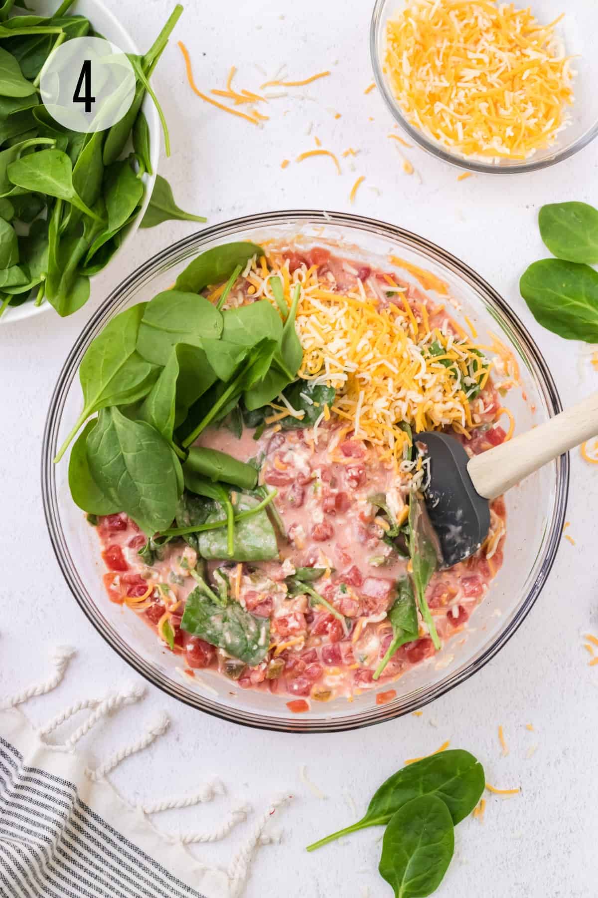 Glass bowl with ingredients for spinach queso dip stirring by grey spatula and bowls of spinach and queso in upper background.