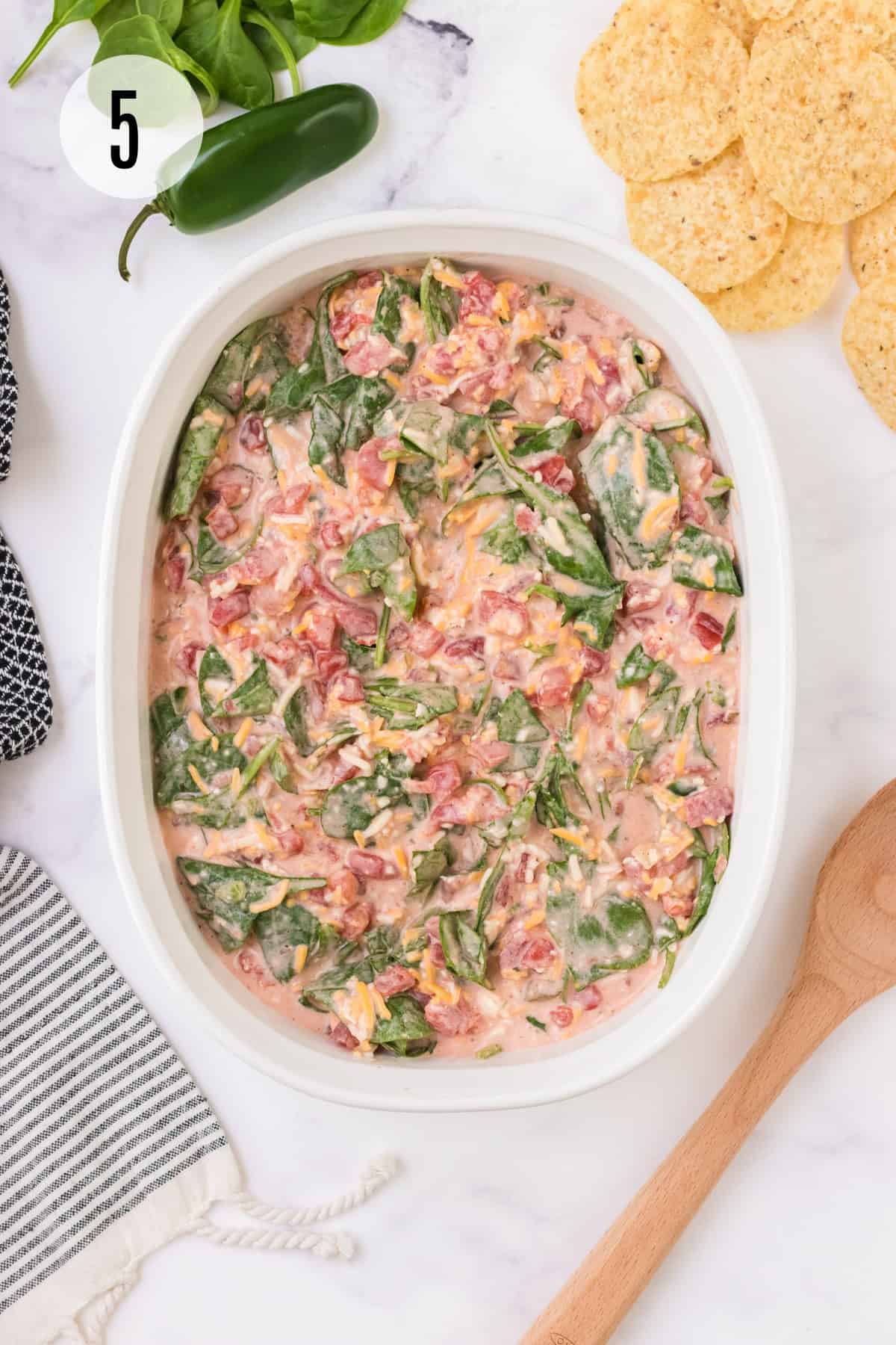 White oval casserole dish with spinach queso dip ready to be baked with tortilla chips and jalapeno in upper background and wooden spoon and black and white linen in lower foreground.