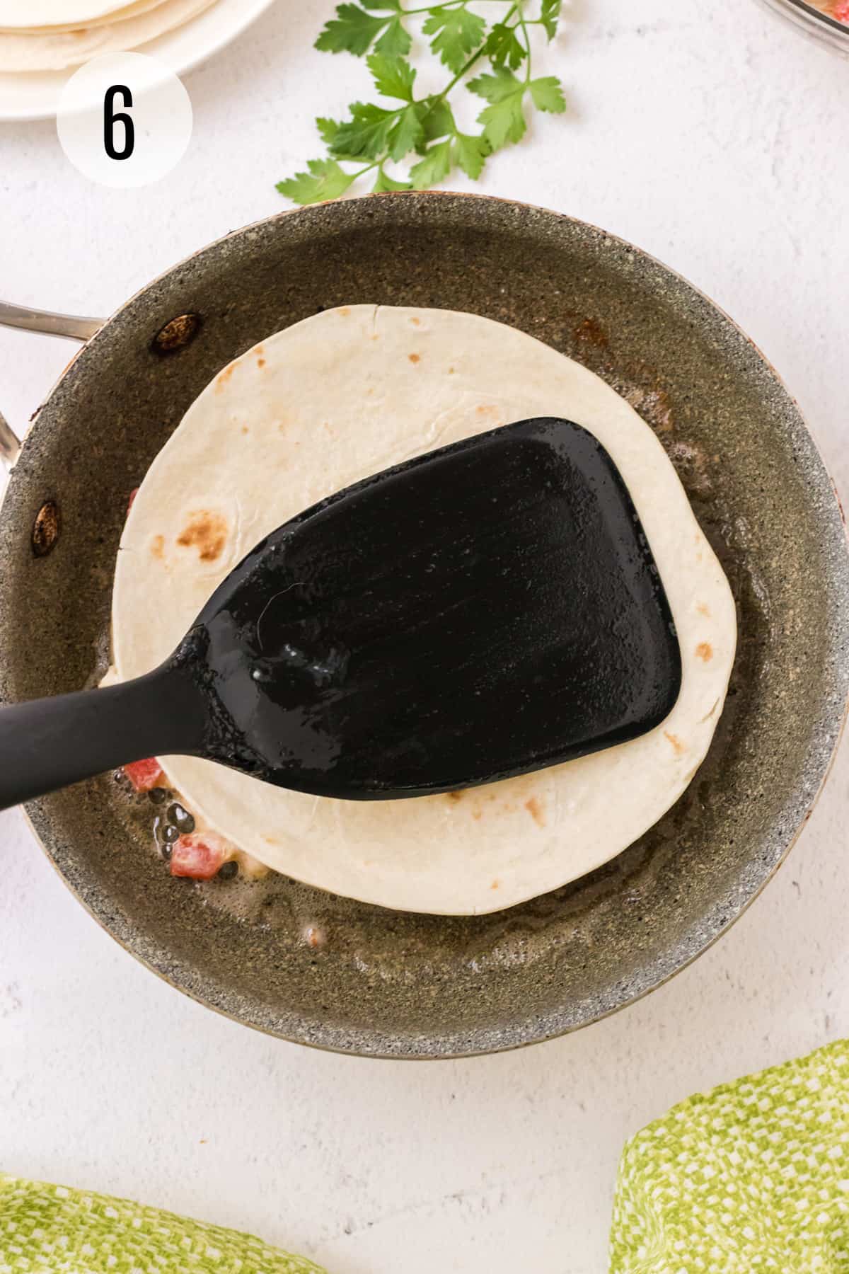 Grey skillet with quesadilla and black spatula pressing together and green parsley in upper background and green linen in lower foreground. 