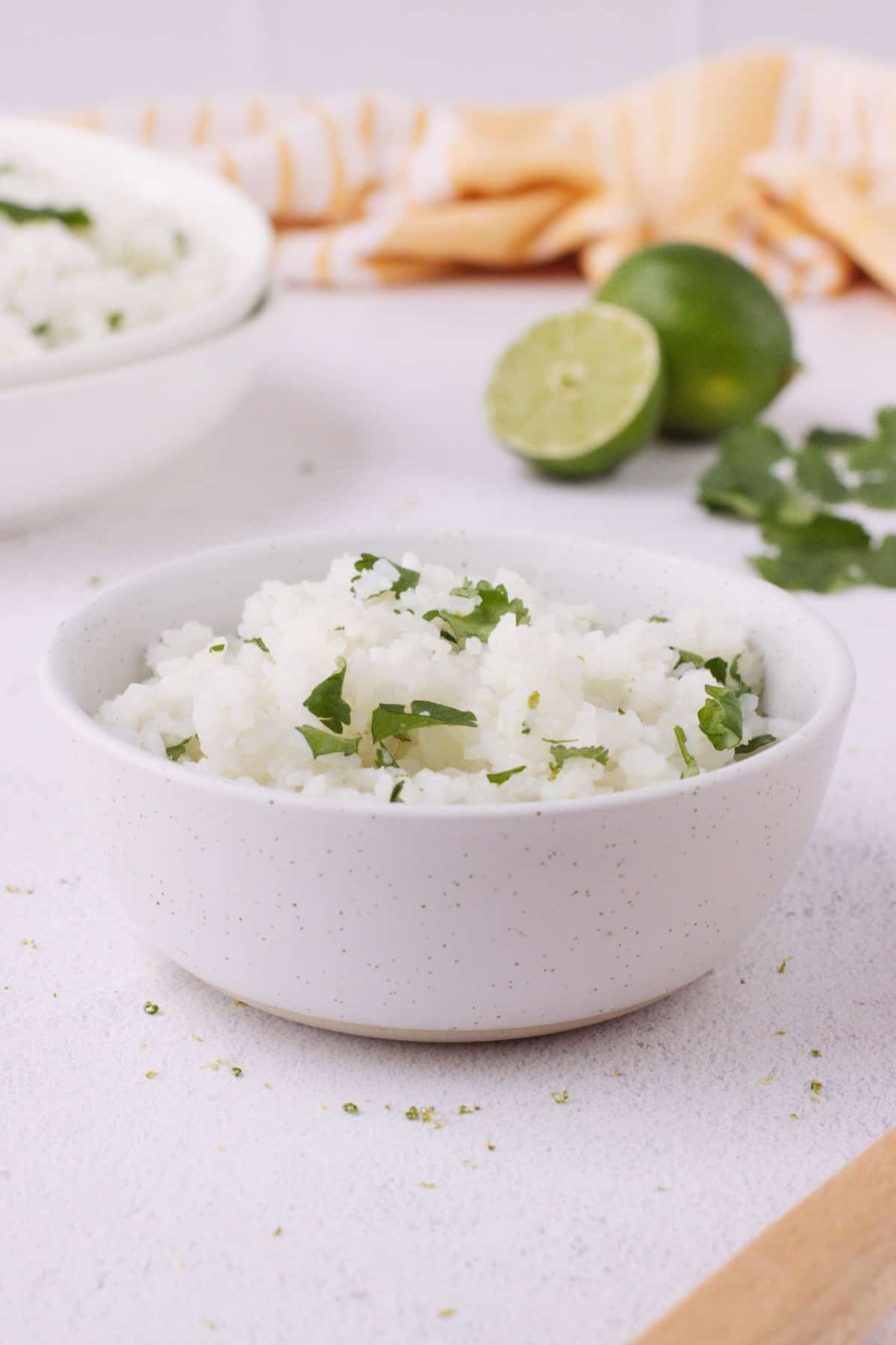 White bowl with white rice and chopped cilantro with cut and whole lime and bowl full of more rice in background with orange linen.
