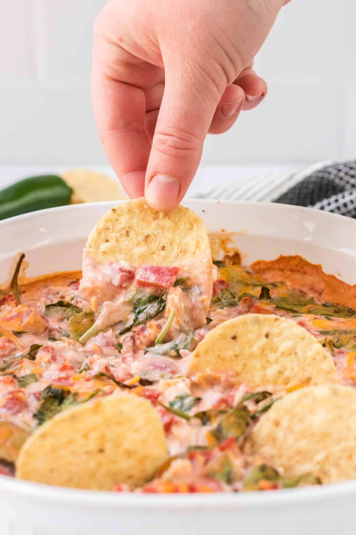 Hand dipping a tortilla chip into spinach queso dip with more chips in foreground placed in dish of dip.