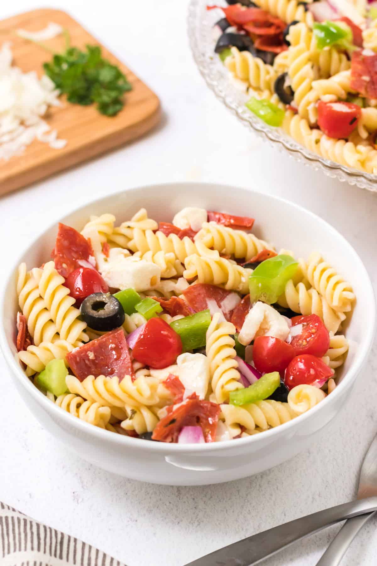 White bowl with Italian pasta salad with tomatoes, green bell peppers, fresh mozzarella, pepperoni, black olives and purple onion with cutting board and large bowl of pasta salad in upper background.
