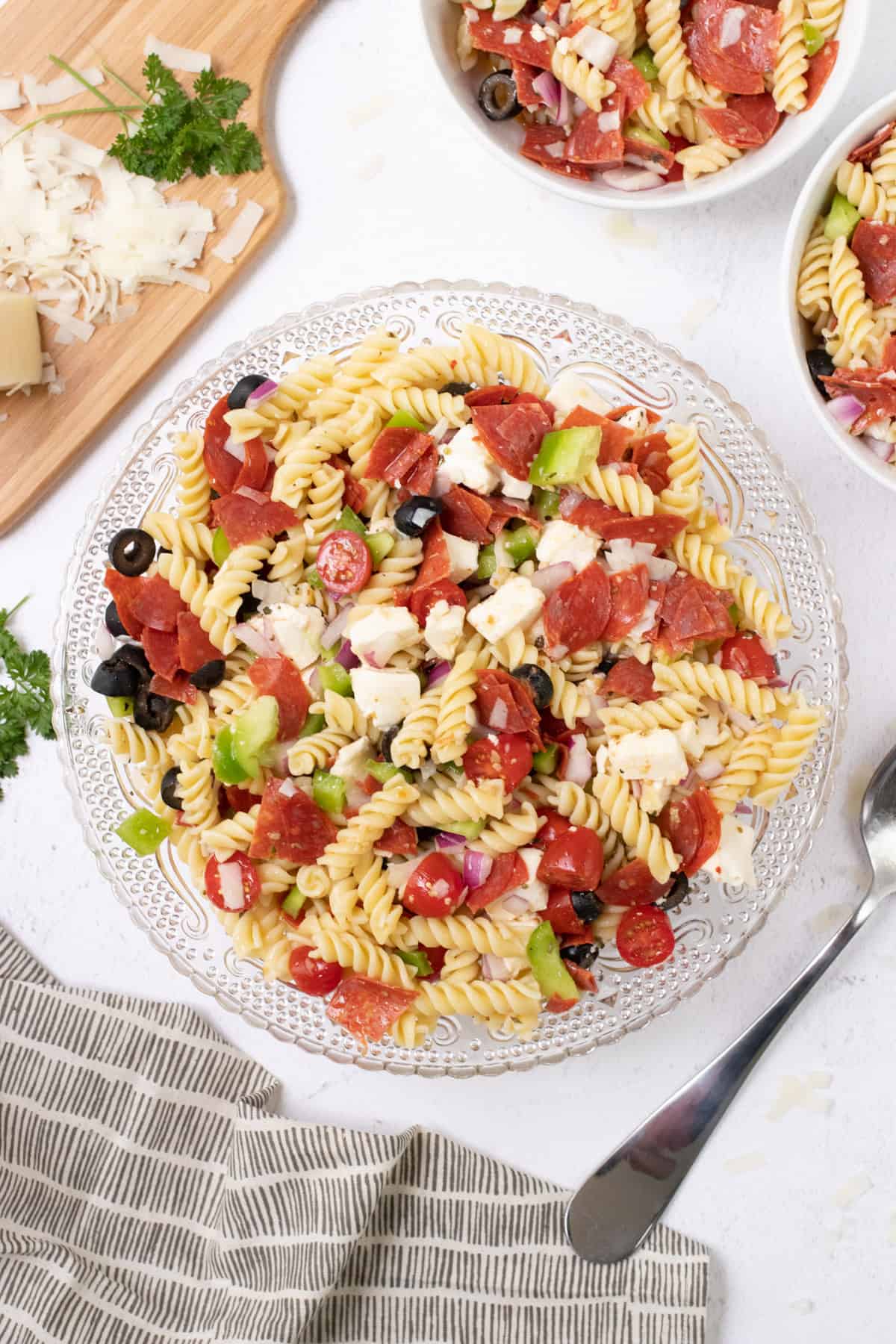 Large glass bowl of Italian pasta salad with pepperoni and fresh vegetables with cutting board of shredded cheese and smaller bowls of pasta salad in upper background.
