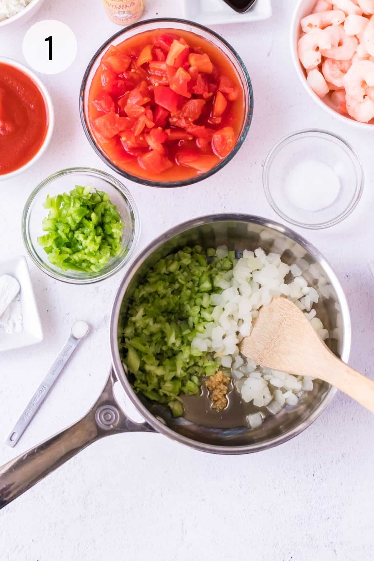 Silver saucepan with chopped celery, onions and garlic in a pan with wooden spoon and other creole sauce ingredients like green bell peppers, diced tomatoes, shrimp and sugar surrounding pan.