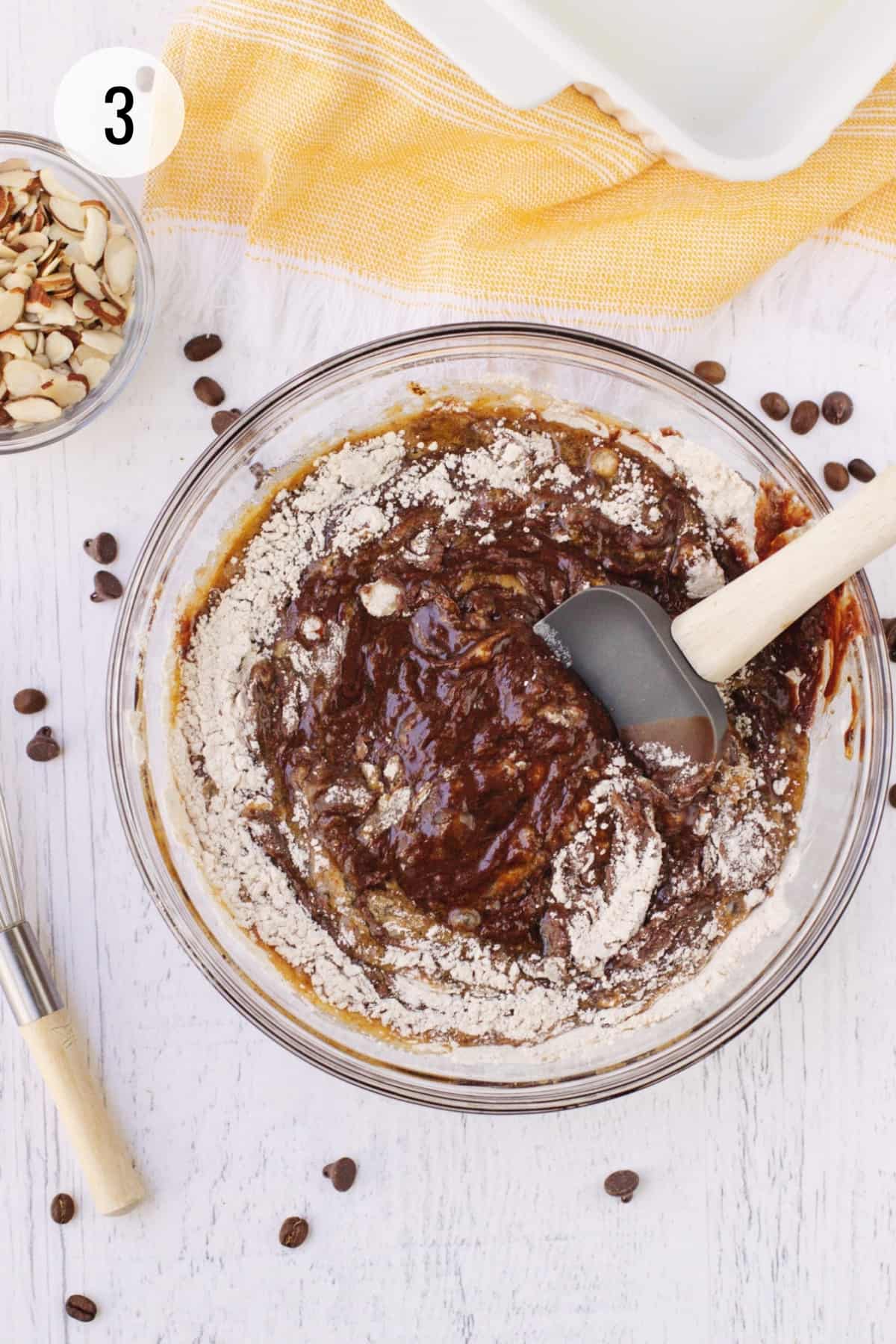 Mexican brownies batter with flour mixture being stirred in by rubber spatula with bowl of sliced almonds and orange linen and white baking dish in upper background. 
