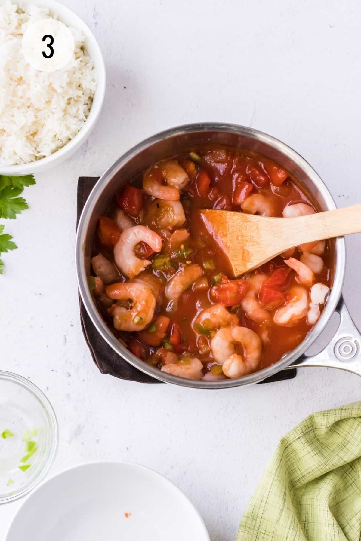 Silver saucepan with shrimp creole sauce stirred by wooden spoon with bowl of white rice in upper left and empty bowls and green linen in lower foreground.