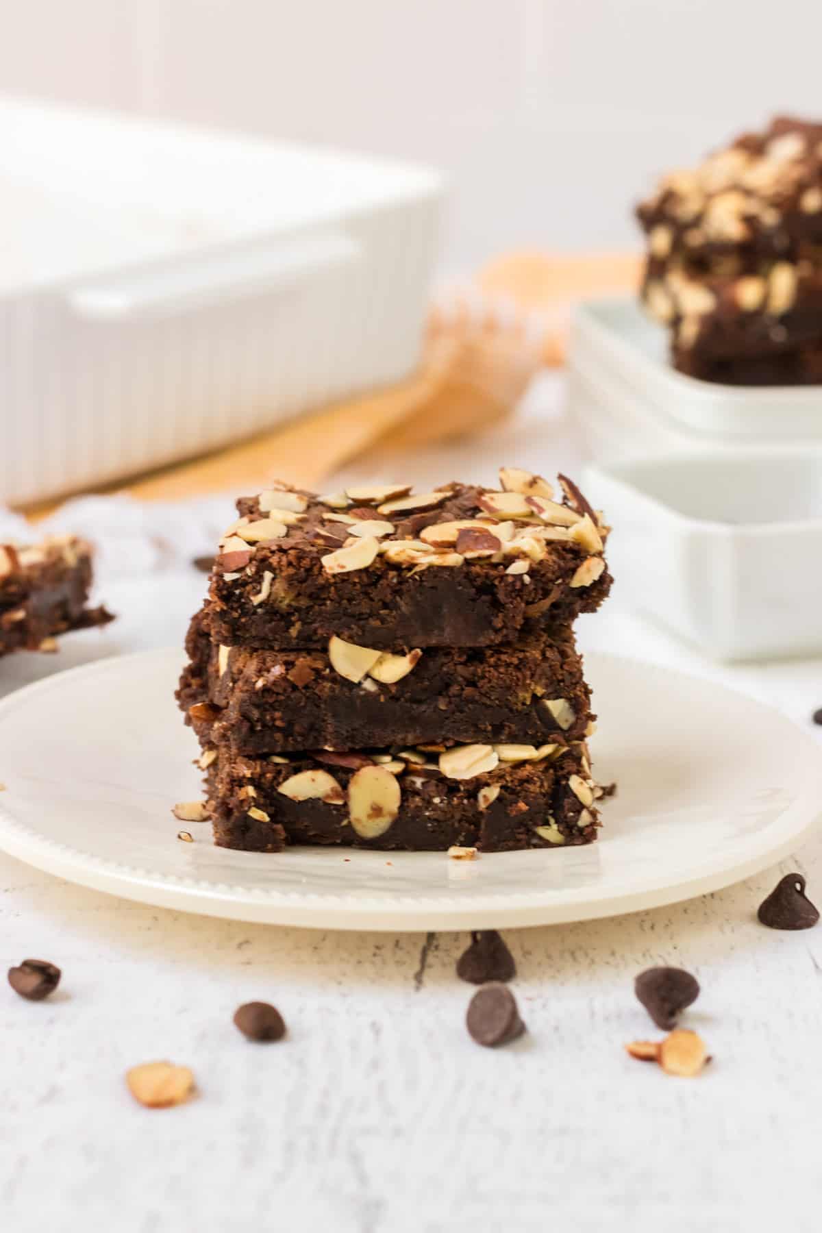 Stack of three Mexican brownies with sliced almonds on a white plate with pan and stack of more brownies in background and chocolate chips sprinkled in foreground. 