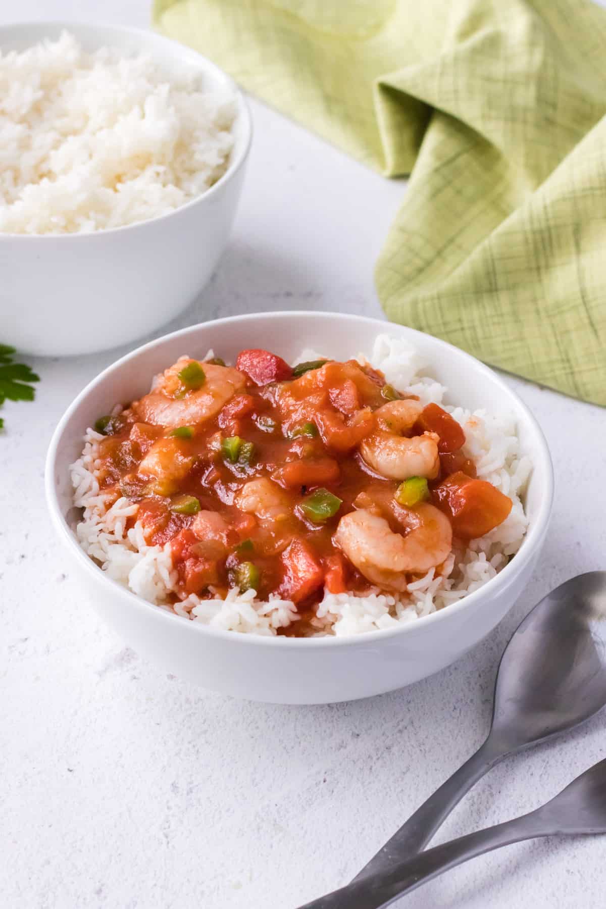 Bowl of shrimp creole sauce over steamed rice with silver spoons in lower right and extra bowl of rice and green linen in upper background.