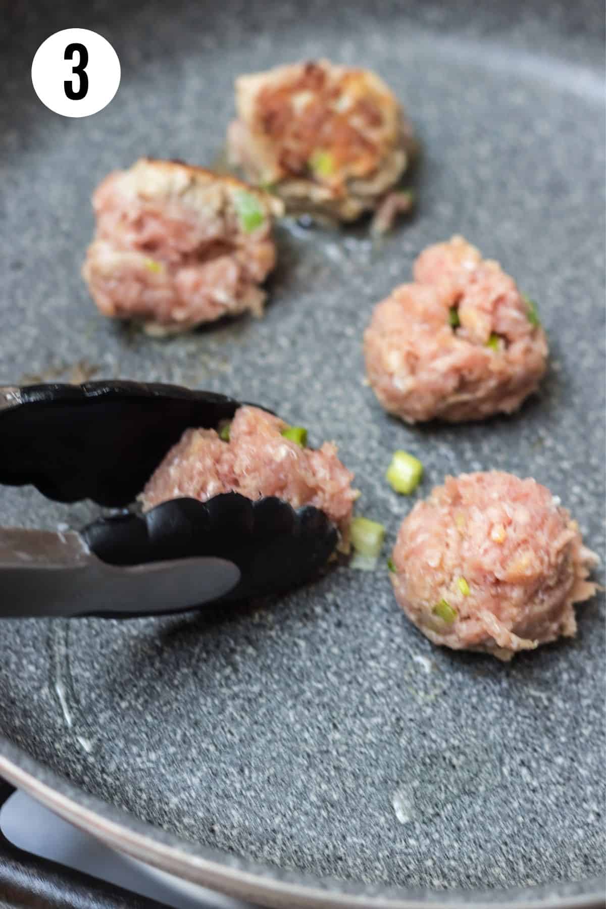 Black tongs turning meatball in a grey skillet with additional meatballs cooking. 