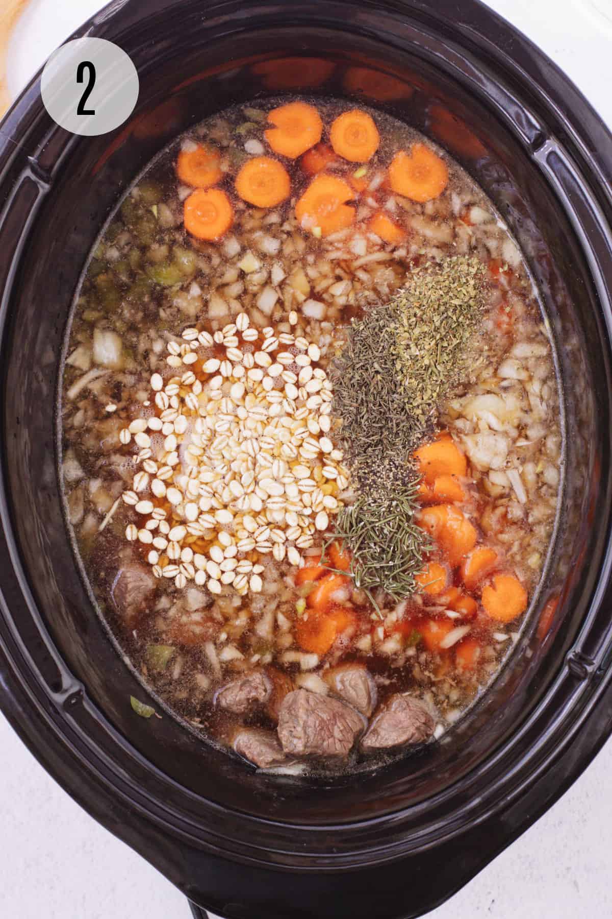 Black crockpot with ingredients for vegetable beef barley soup including chopped carrots, dried herbs, beef and barley. 