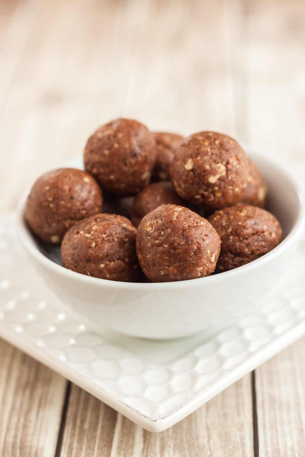 White bowl of chocolate peanut butter protein balls on square white plate. 