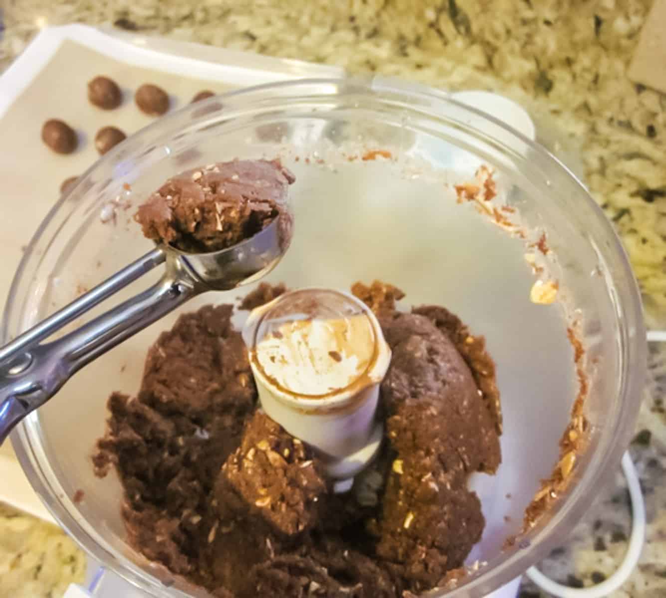 Food processor bowl with chocolate peanut butter protein ball mixture and metal scoop scooping out balls. 