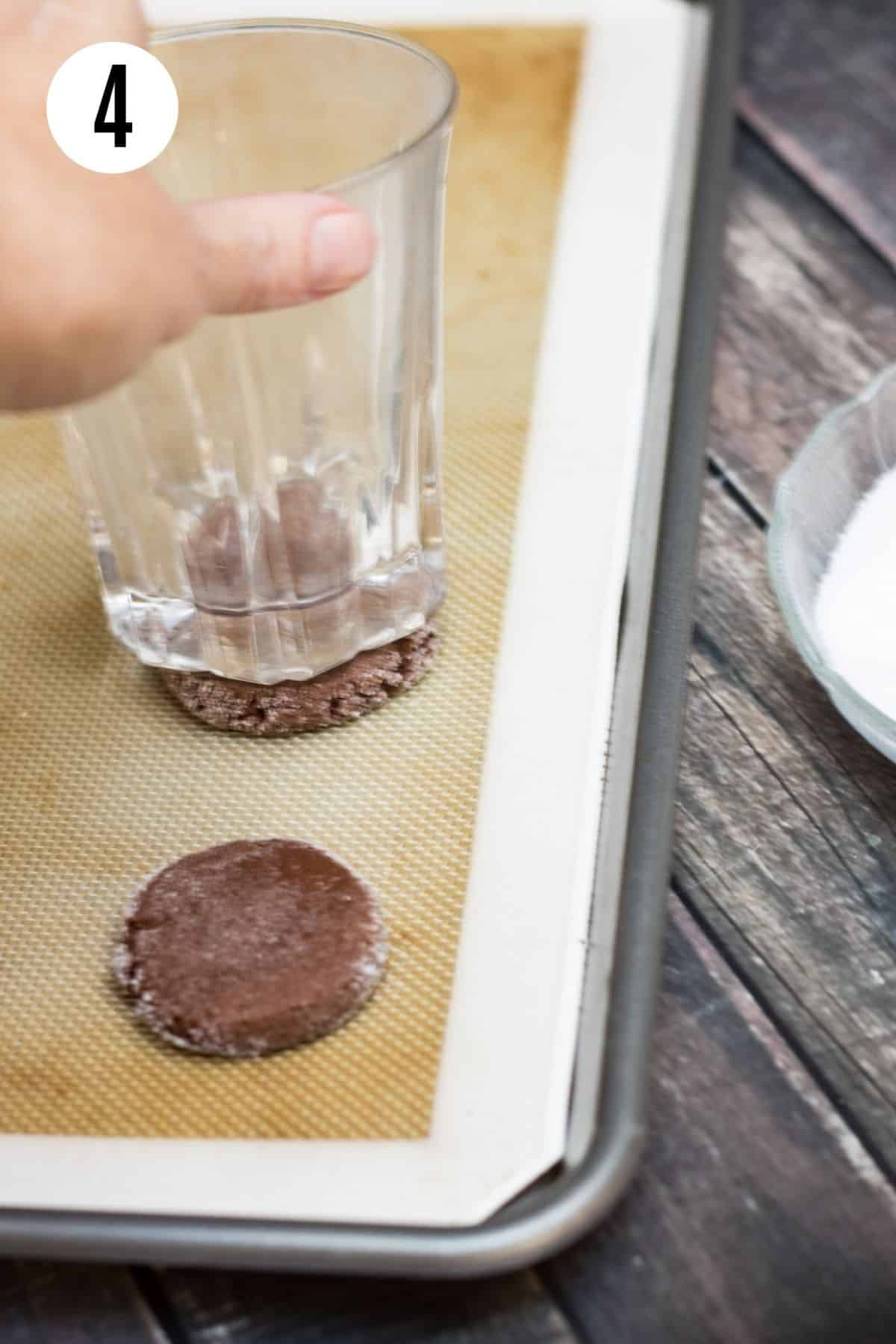 Clear drinking glass pressing chocolate cookie dough flattening on a baking sheet lined with silicone baking mat. 