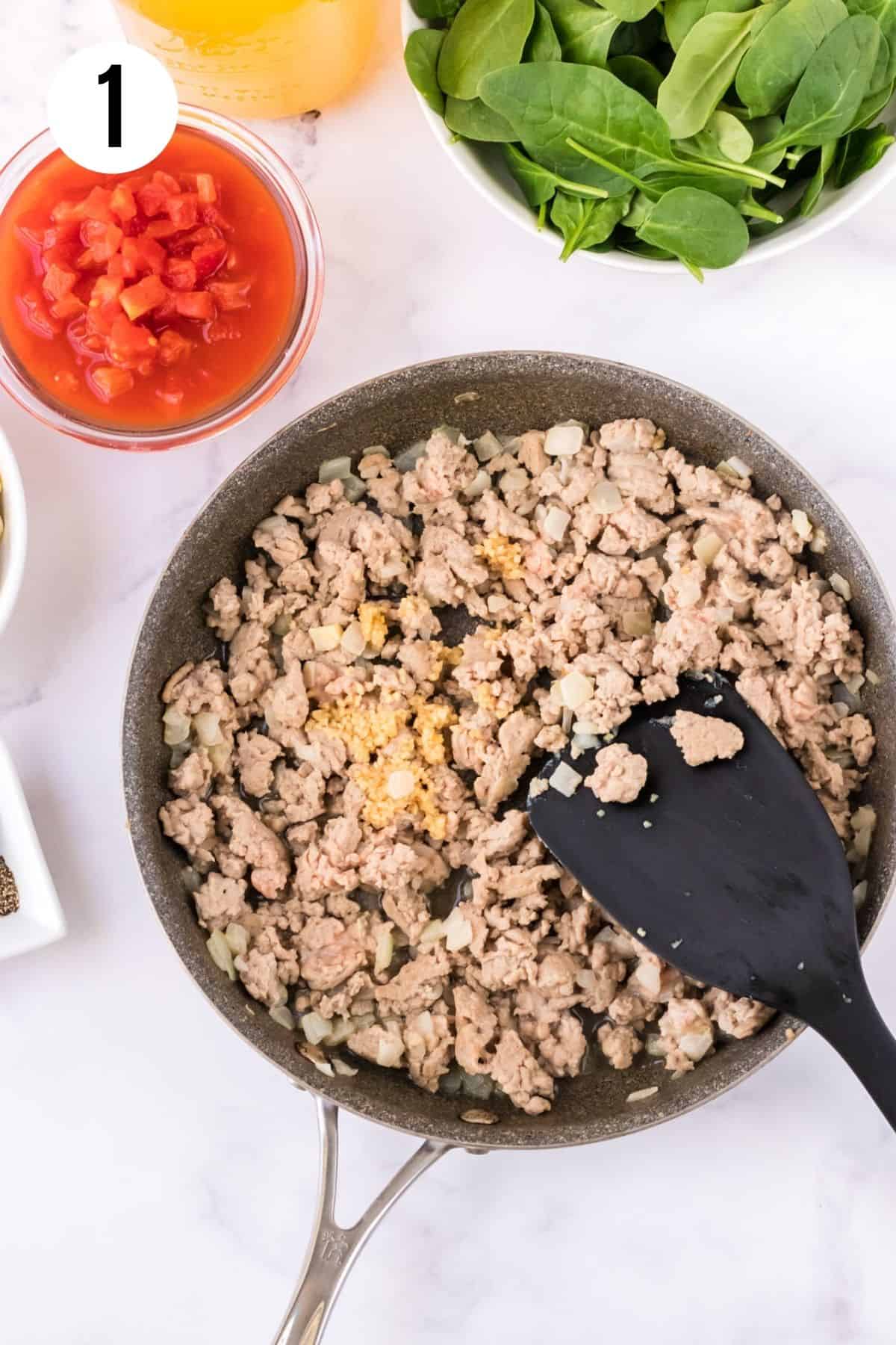 Skillet with browning ground turkey sausage and minced garlic and onions stirred by black spatula with bowls of diced tomatoes, baby spinach and chicken broth in upper background.