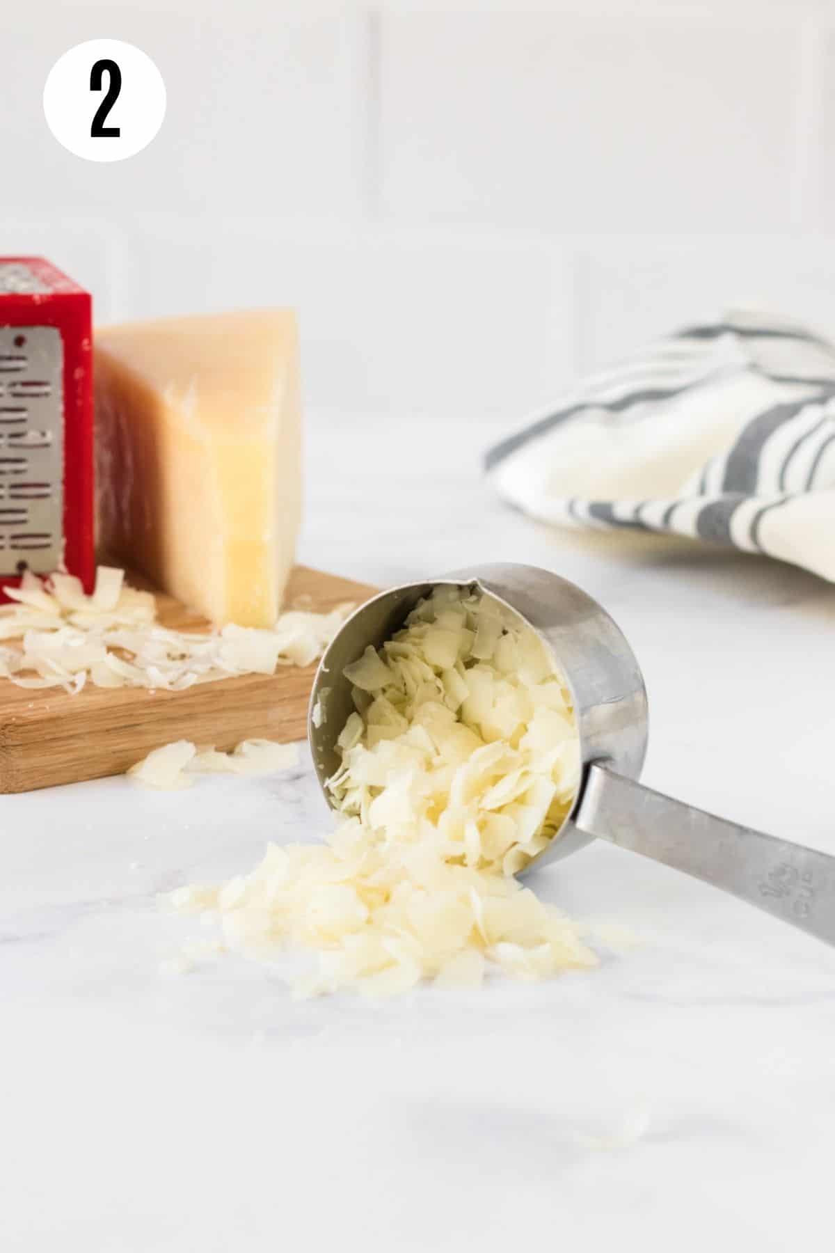 Metal measuring cup with large shreds of Parmesan cheese spilling out and chunk of cheese and red box grater and black and white linen in background.
