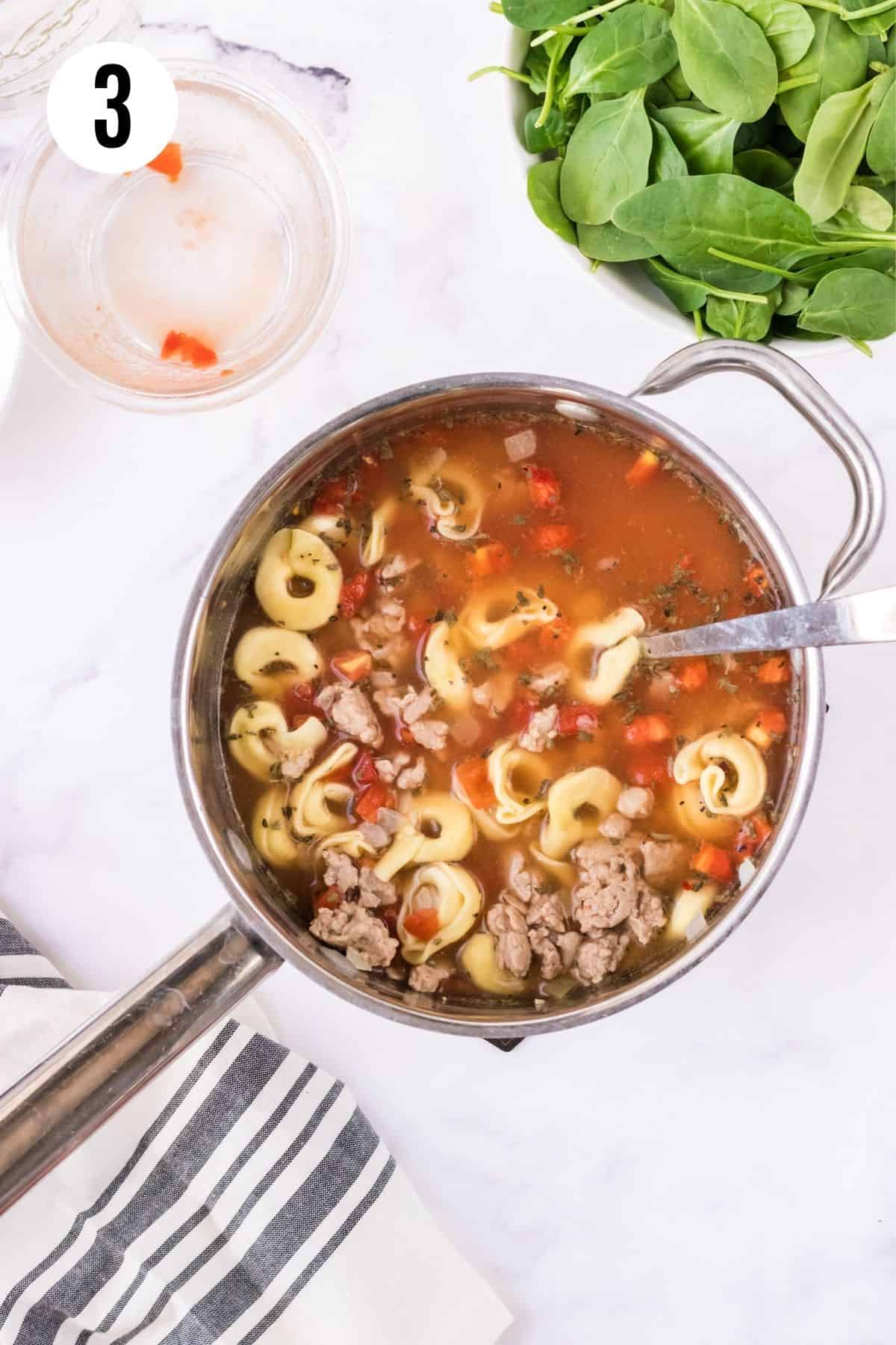 Top view of saucepan of Italian sausage and tortellini soup with bowl of baby spinach in upper right, empty glass bowl in upper left and black and white linen in lower left.