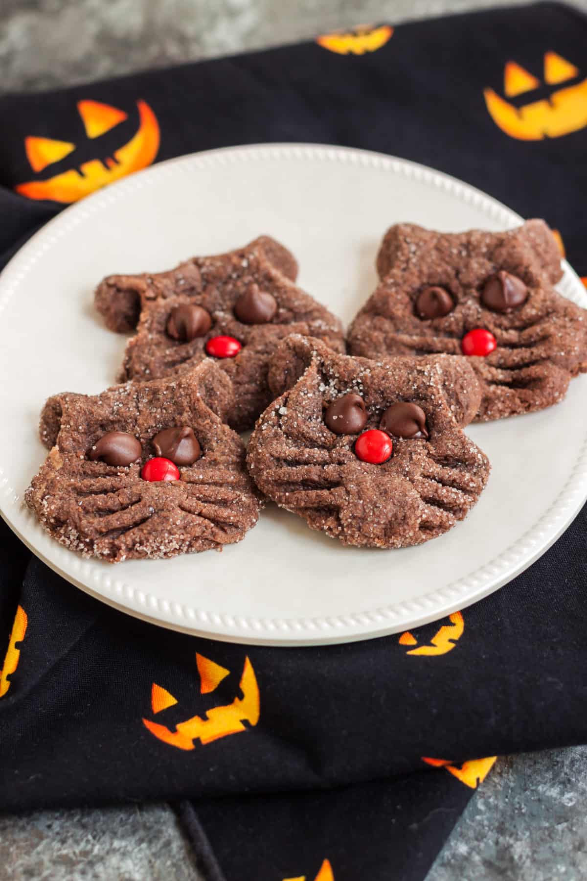 White plate of chocolate cookies shaped like a black cat face with chocolate chip eyes and red candy nose on a black and yellow jack-o-lantern napkin background. 
