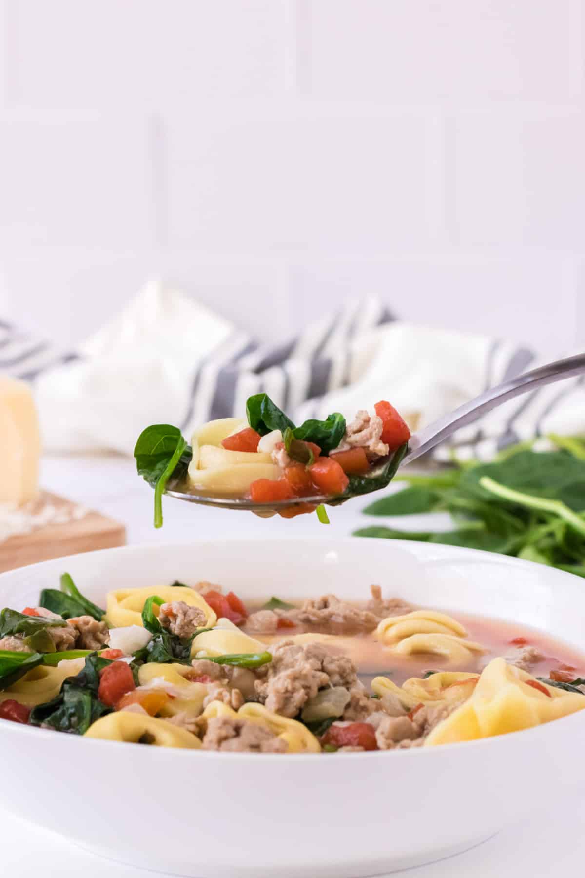 White bowl of Italian sausage tortellini soup with spinach and tomatoes and spoonful of soup on silver spoon with black and white linen and baby spinach in background.